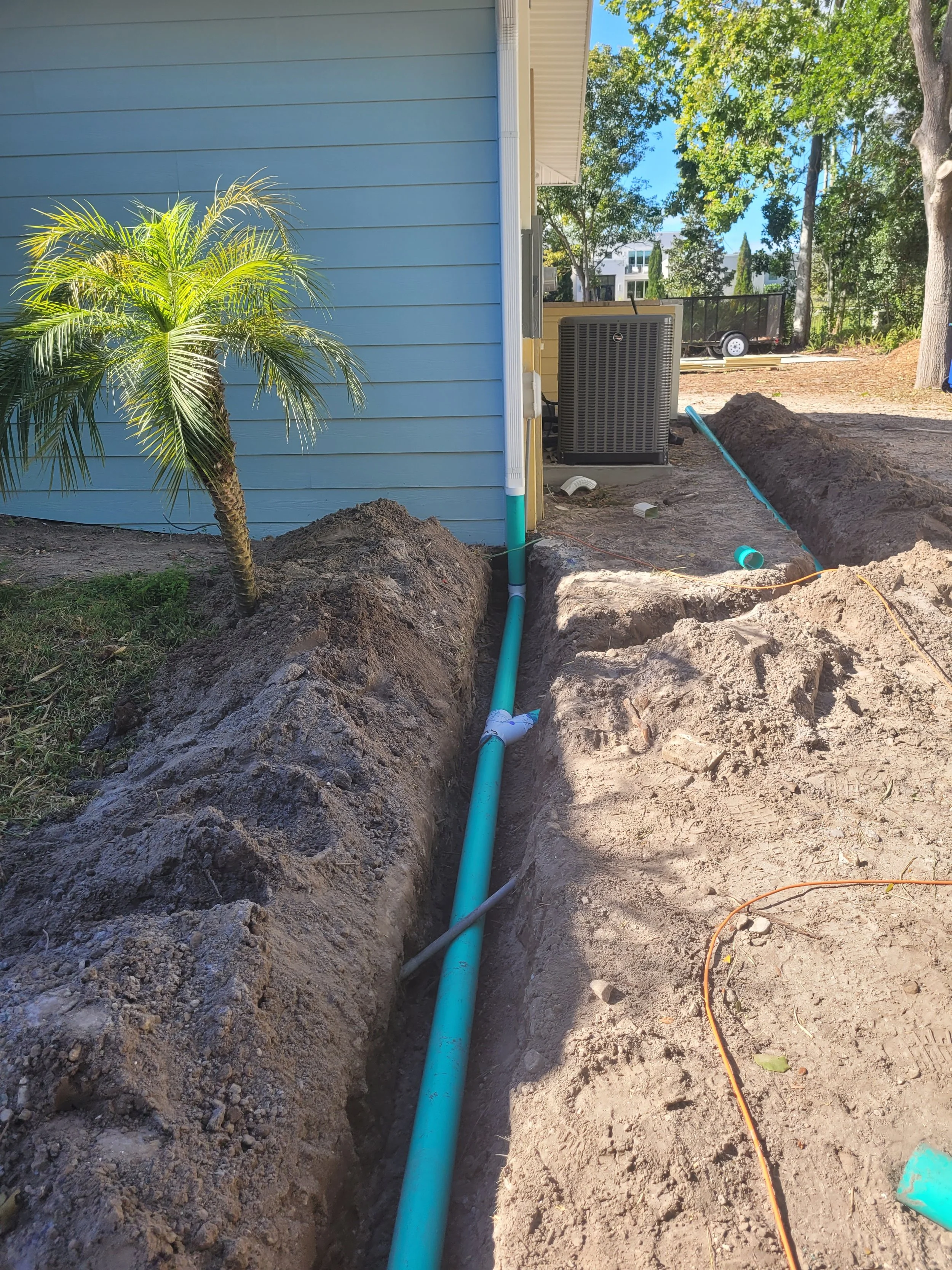 A trench dug along the side of a house with a new blue pipe installed inside it, and a small palm tree nearby on the left, in a residential yard with trees and a trailer in the background.