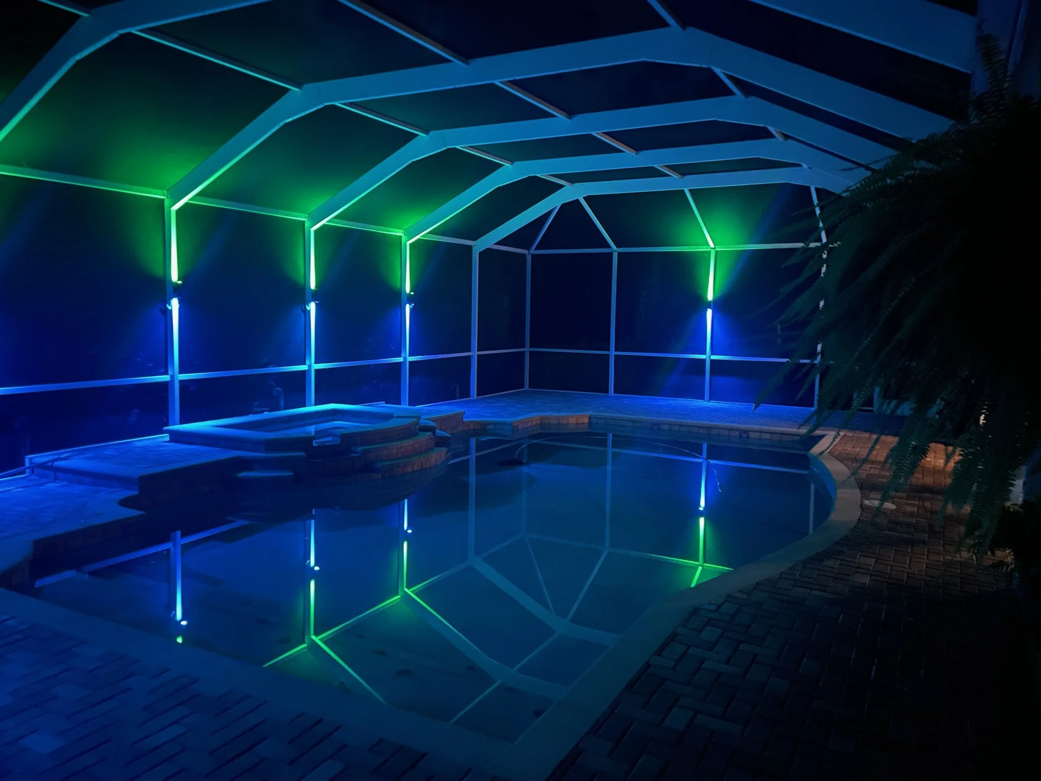 Indoor pool area at night with blue and green LED lights, enclosed by a screened structure, with brick steps leading into the pool and a brick deck surrounding it.