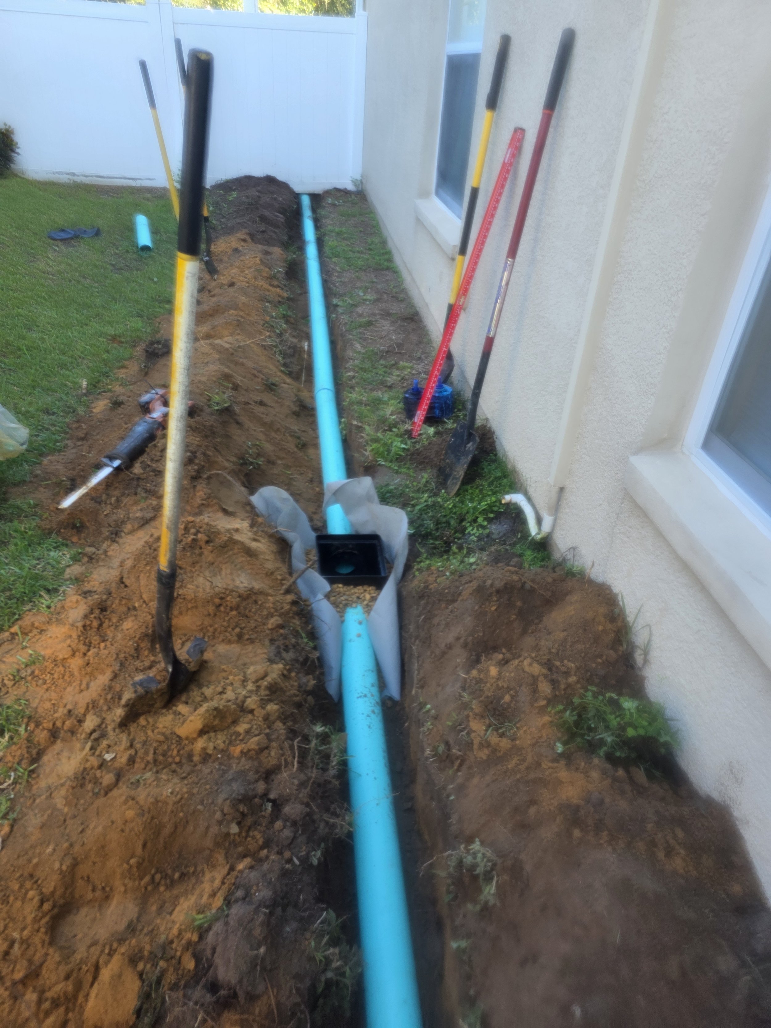 A trench dug alongside a house with a blue underground pipe installed, surrounded by shovels and tools.