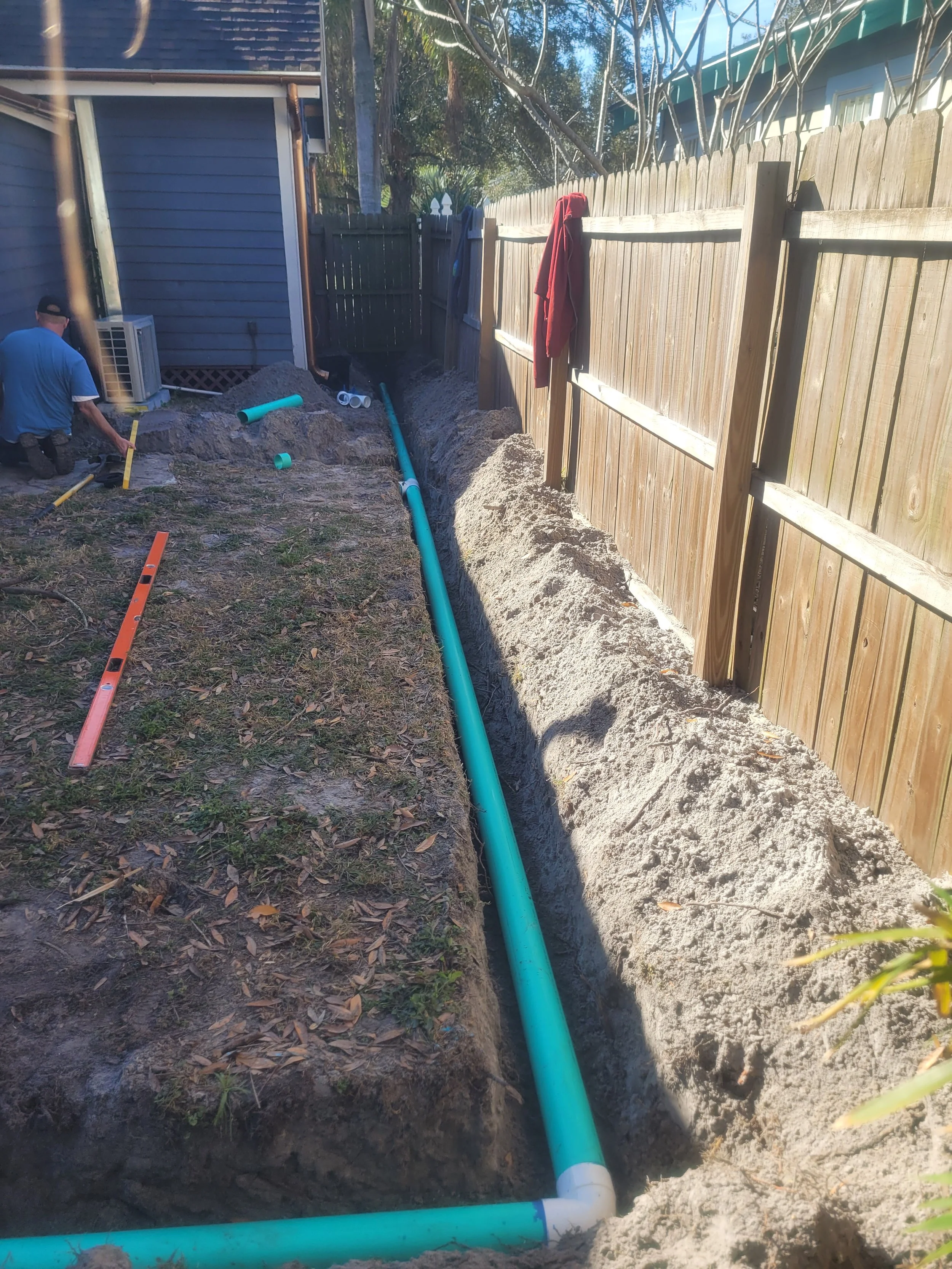 Construction worker installing a blue underground pipe in a trench alongside a wooden fence, with tools and additional pipes nearby.