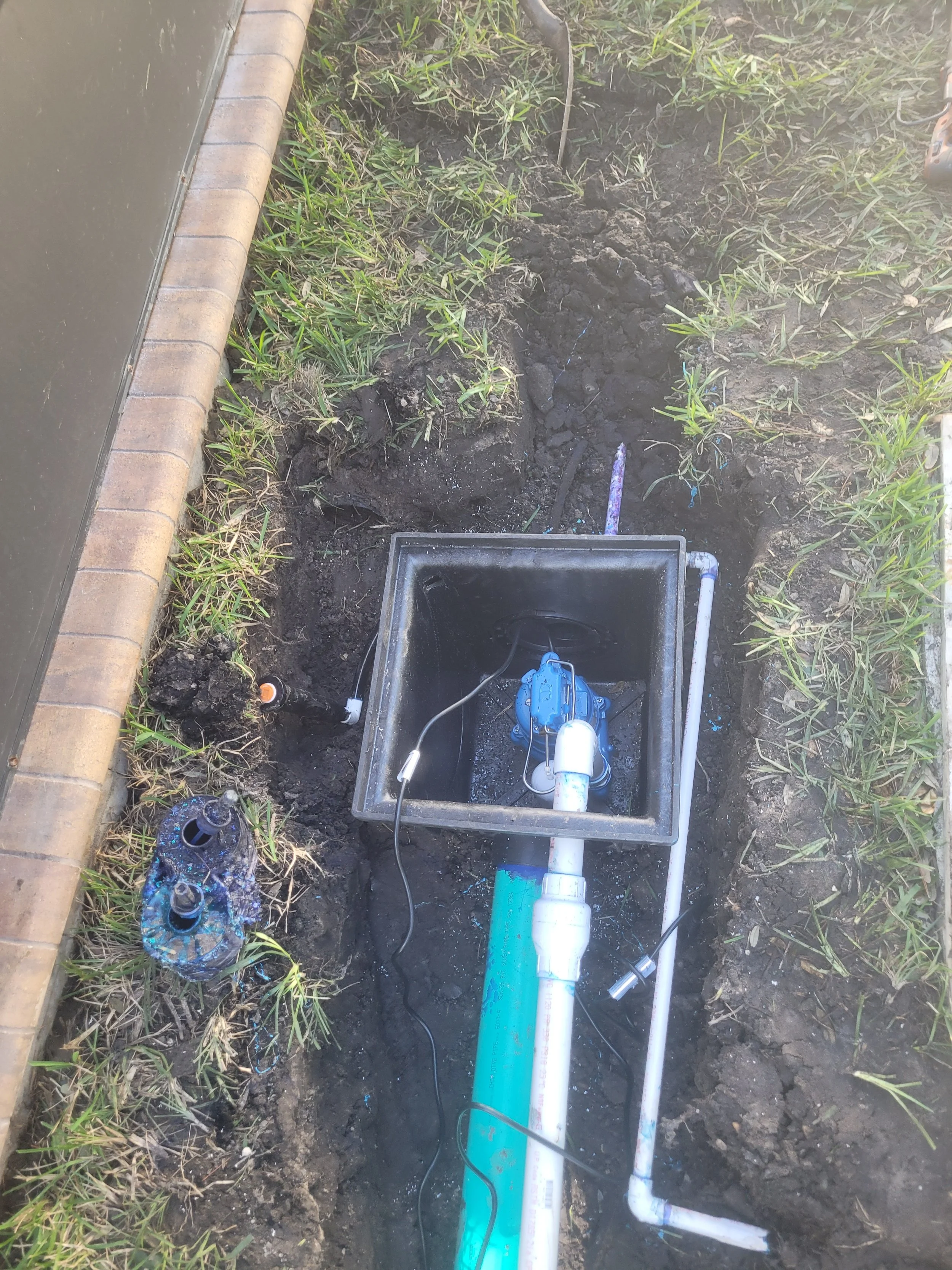 An underground water pump installation in a trench with pipes, an electric cable, and a water valve. The trench is next to a brick border and area with grass.