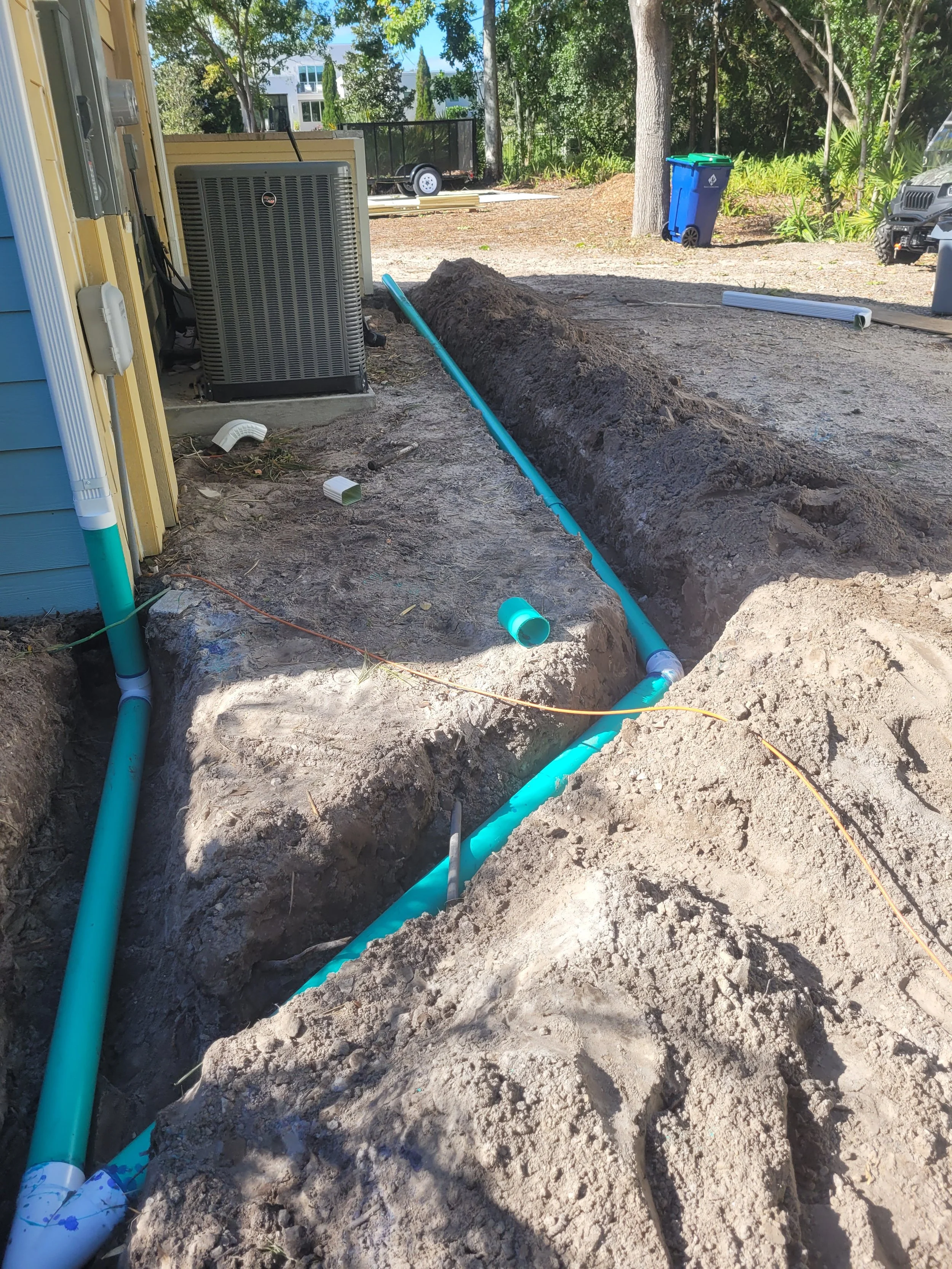 Installation of underground plumbing pipes outside a house, with trench dug for pipes, and various construction tools and equipment visible in the background.