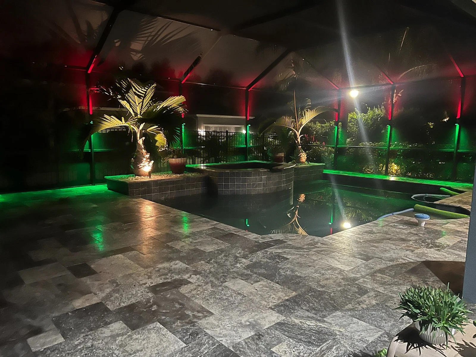 Night view of an outdoor enclosed swimming pool area with red and green lights, potted plants, and a tiled hot tub.