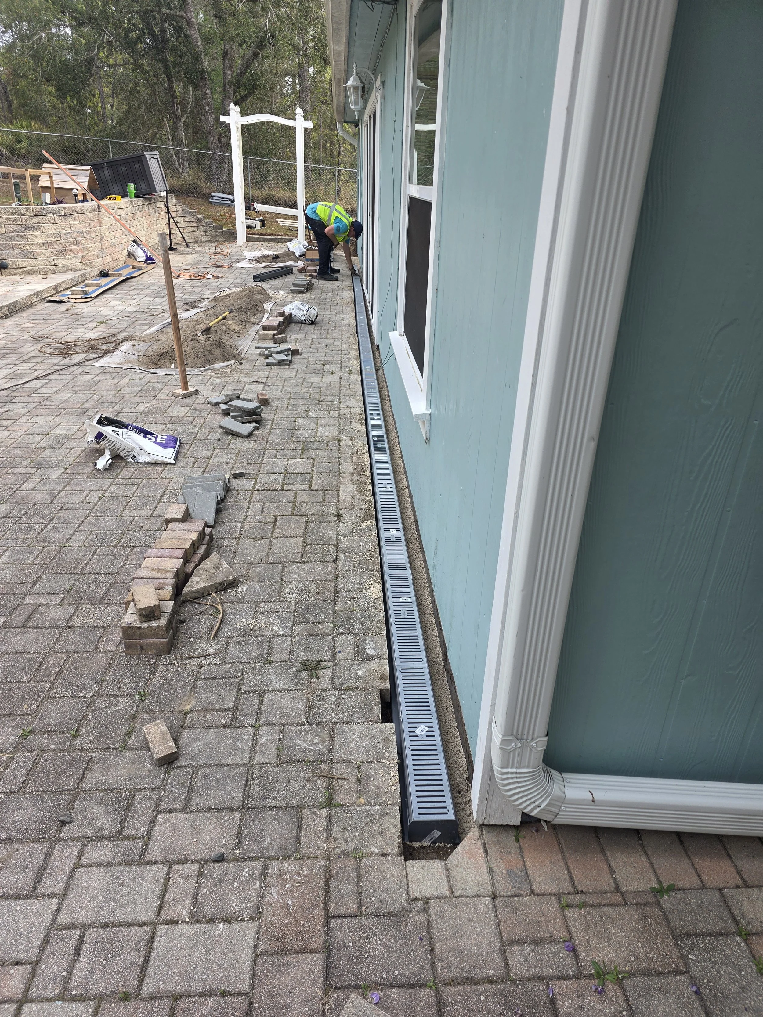 Person working on installing or repairing a drainage system along the foundation of a light blue house with a brick patio. The worker is wearing a safety vest and is bent over, doing construction work. There are building materials and tools scattered