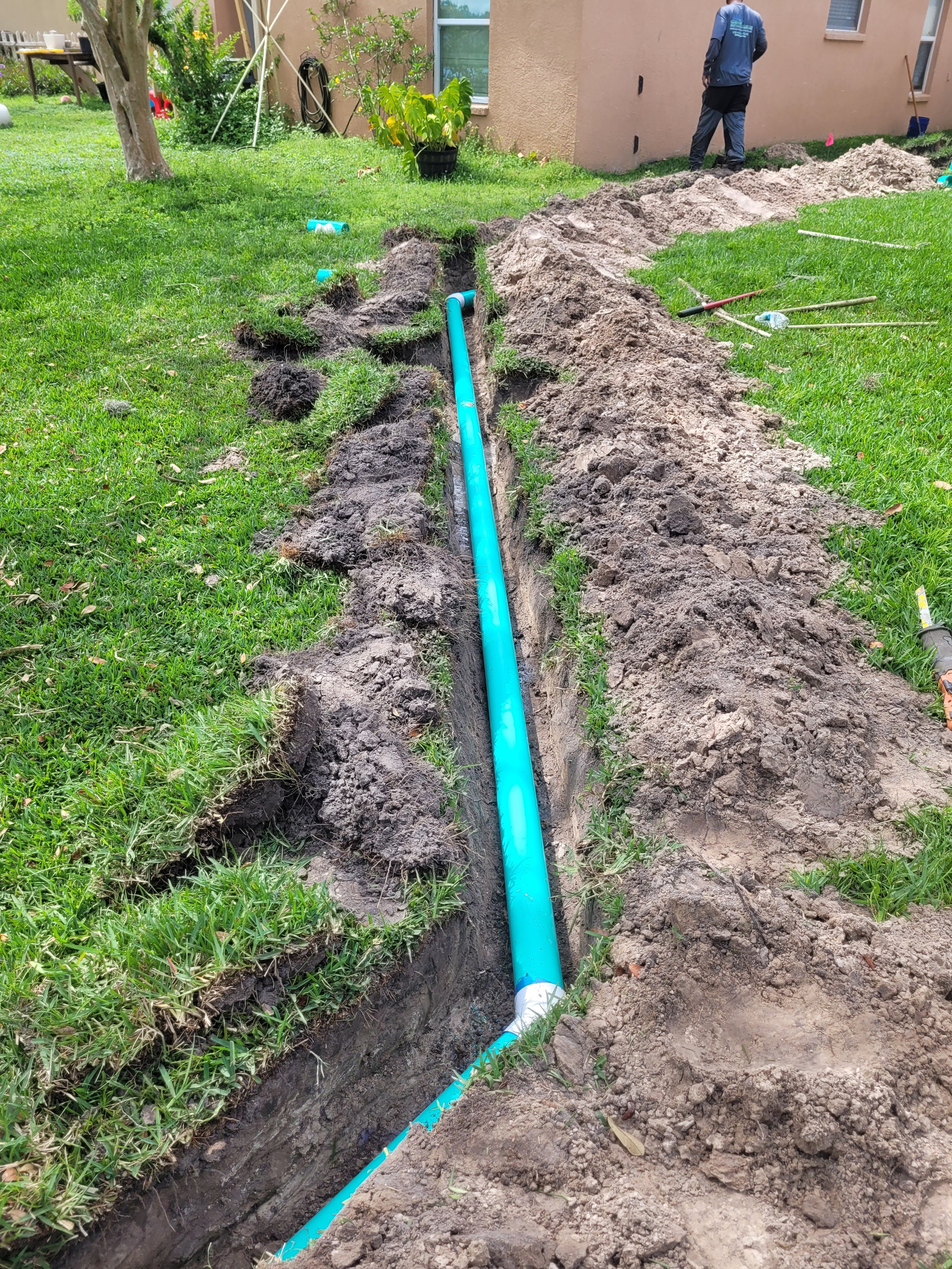 A trench with a blue pipe being installed in a grassy backyard, with tools and a worker in the background.