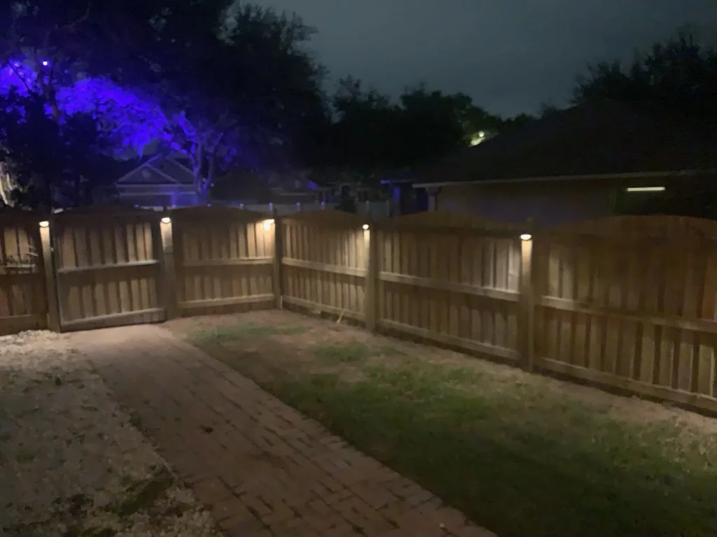 Fenced backyard at night with outdoor lighting and trees illuminated by purple lights.