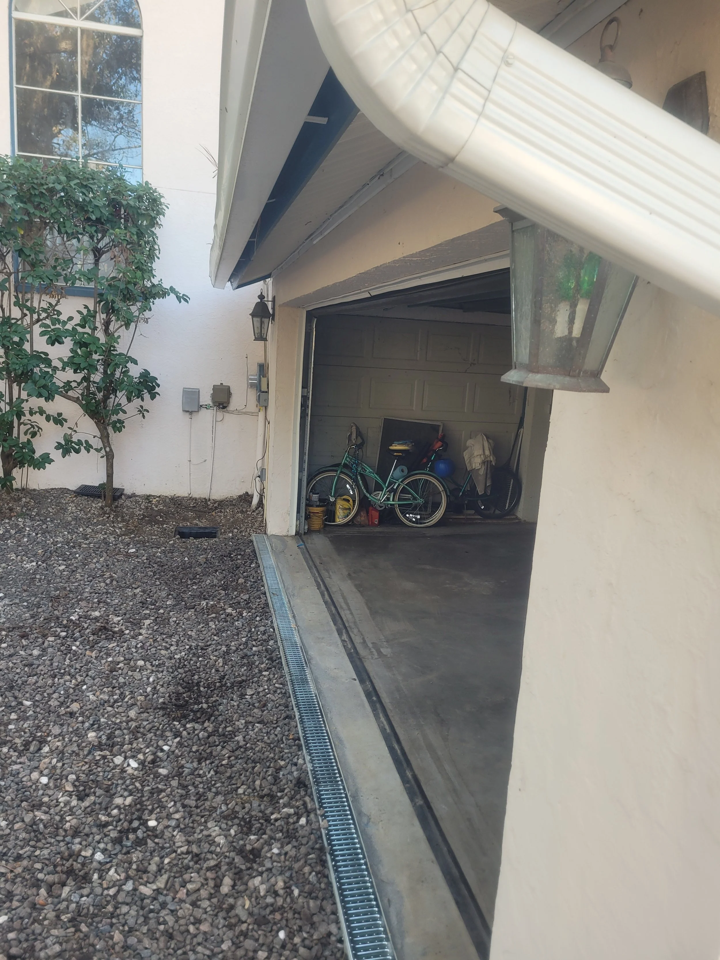 View of a garage with a bicycle, a blue ball, and other items inside, next to a gravel area and a house with a window and a small tree.