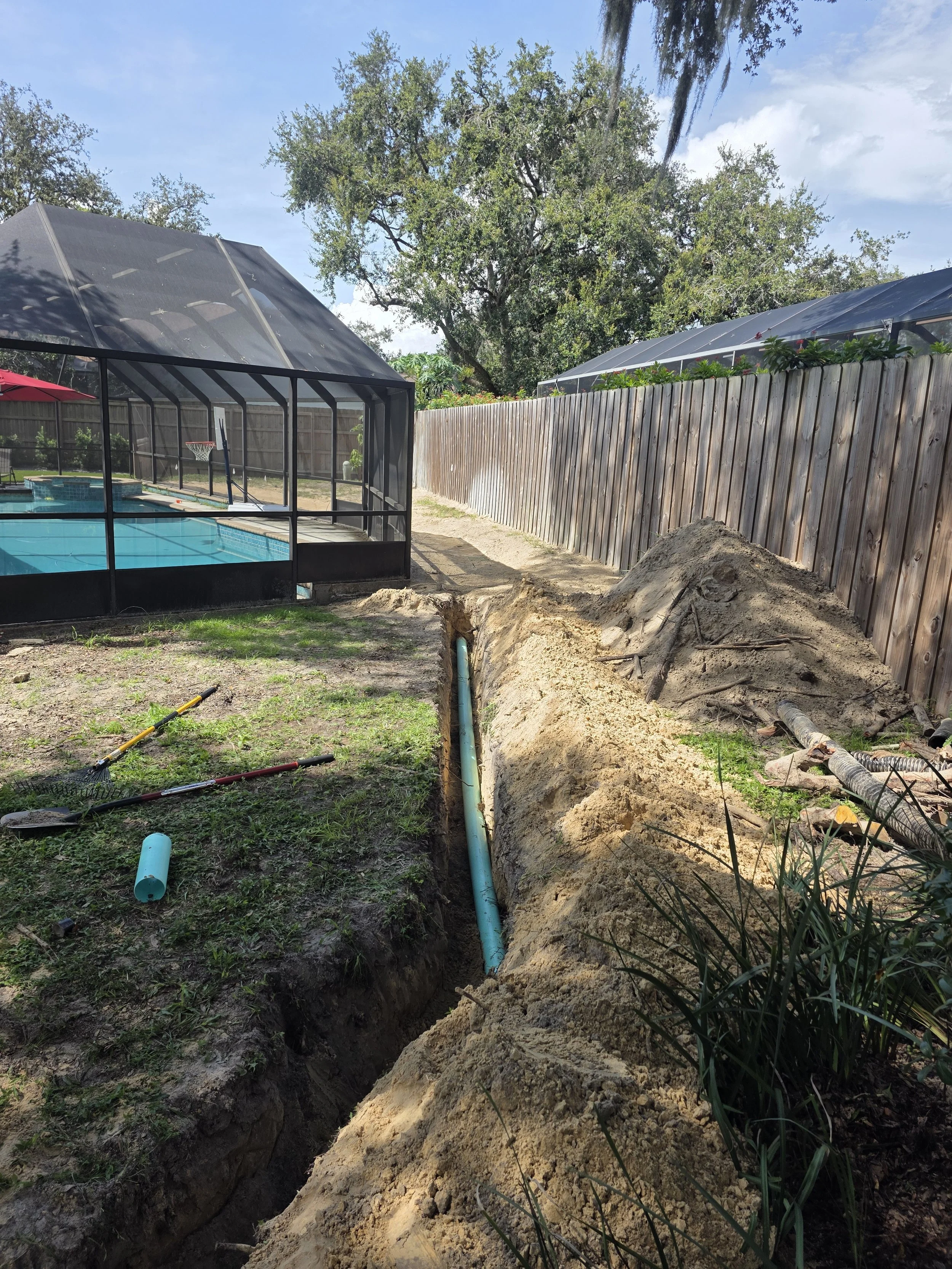 A backyard with a fenced pool enclosure, a swimming pool, and a trench with exposed pipes being installed for plumbing work.