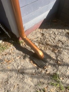 Close-up of a copper downspout directing water into a black drainage grate on sandy ground outside a building.