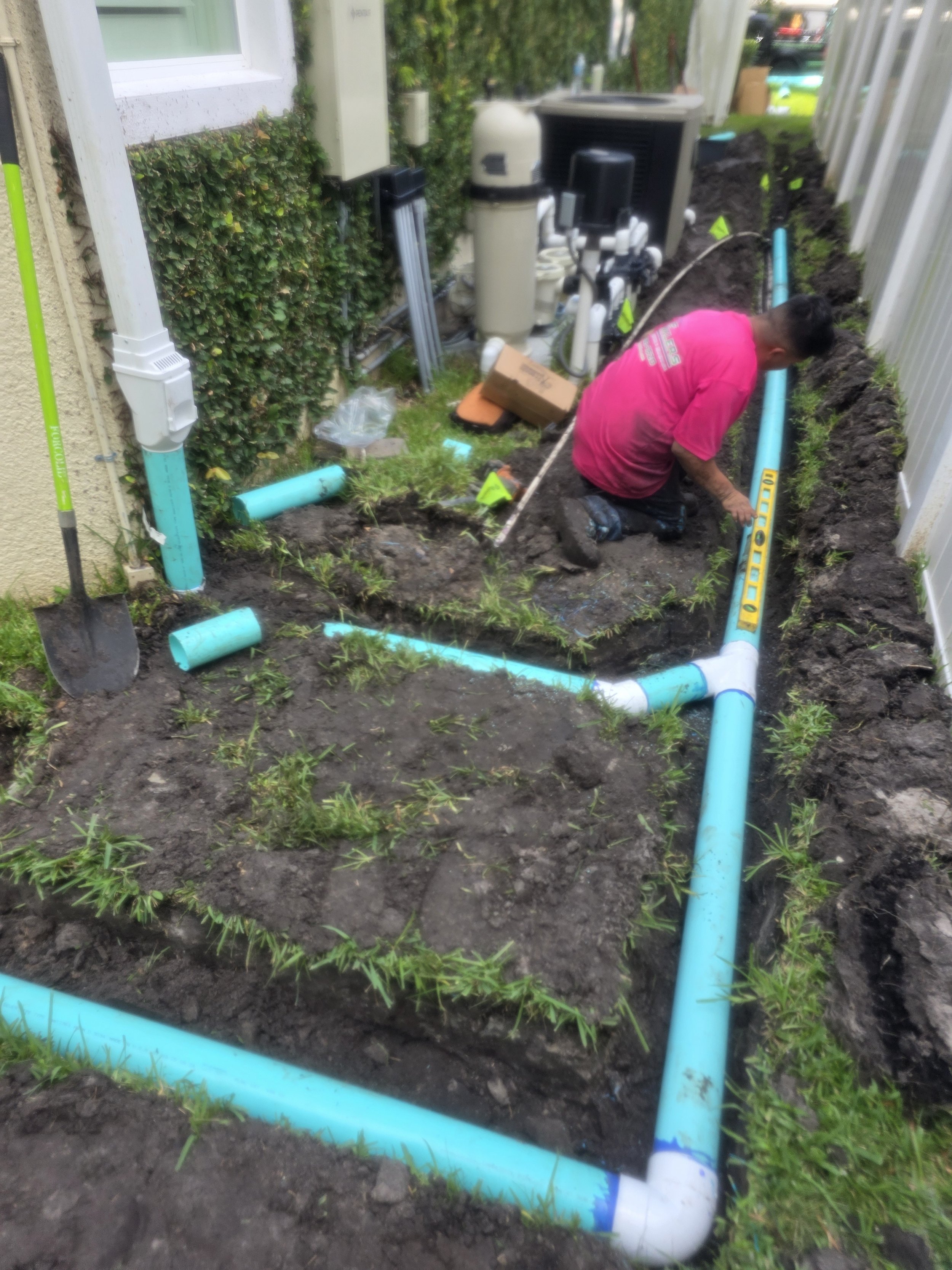 A man working on installing a pool piping system in a backyard, with blue PVC pipes laid out in a rectangular shape along the ground, surrounded by open soil and small plants.