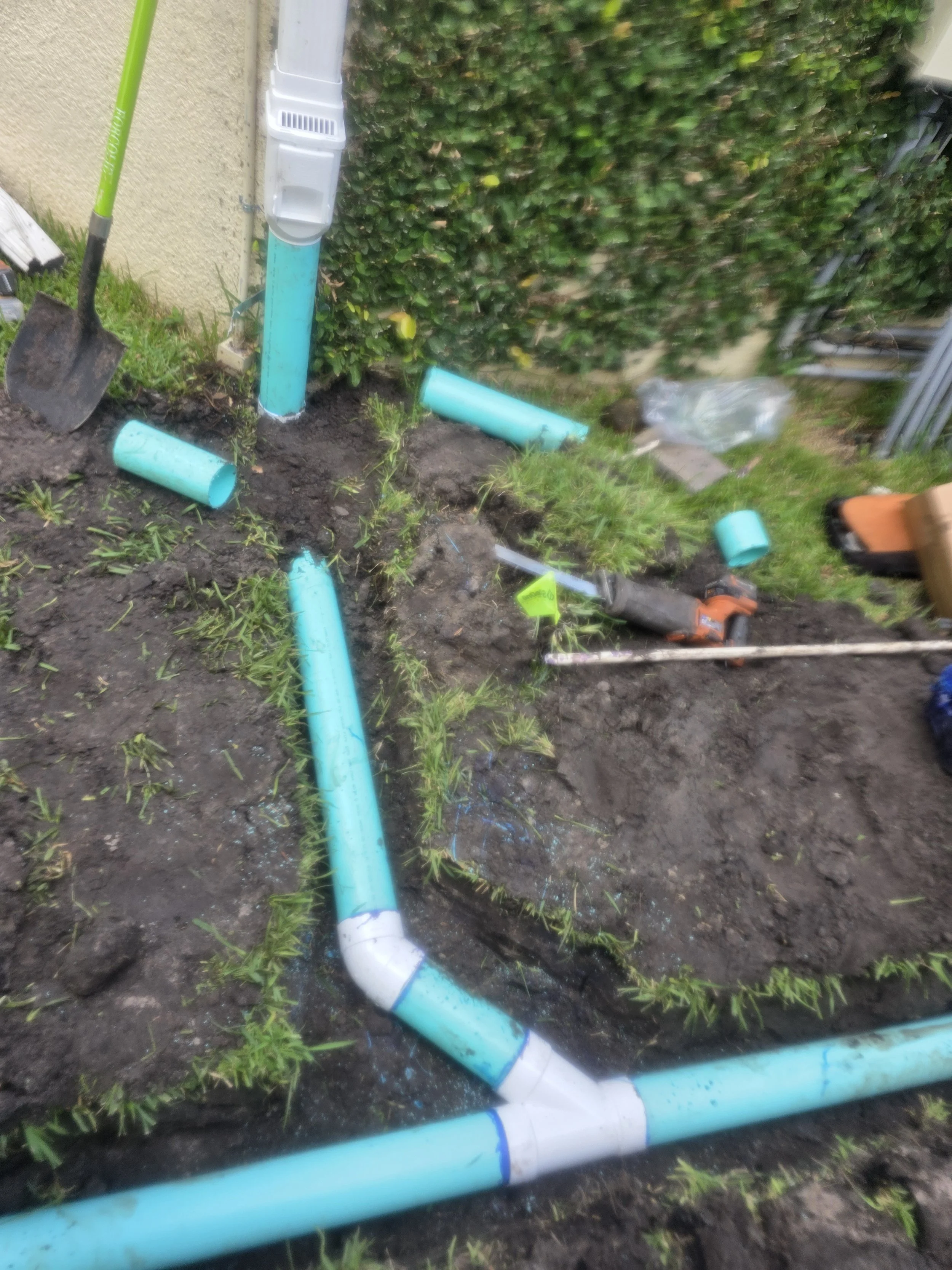 Installing or repairing underground plumbing with blue PVC pipes in a garden area near a wall and hedge, with tools like a shovel and a saw visible.