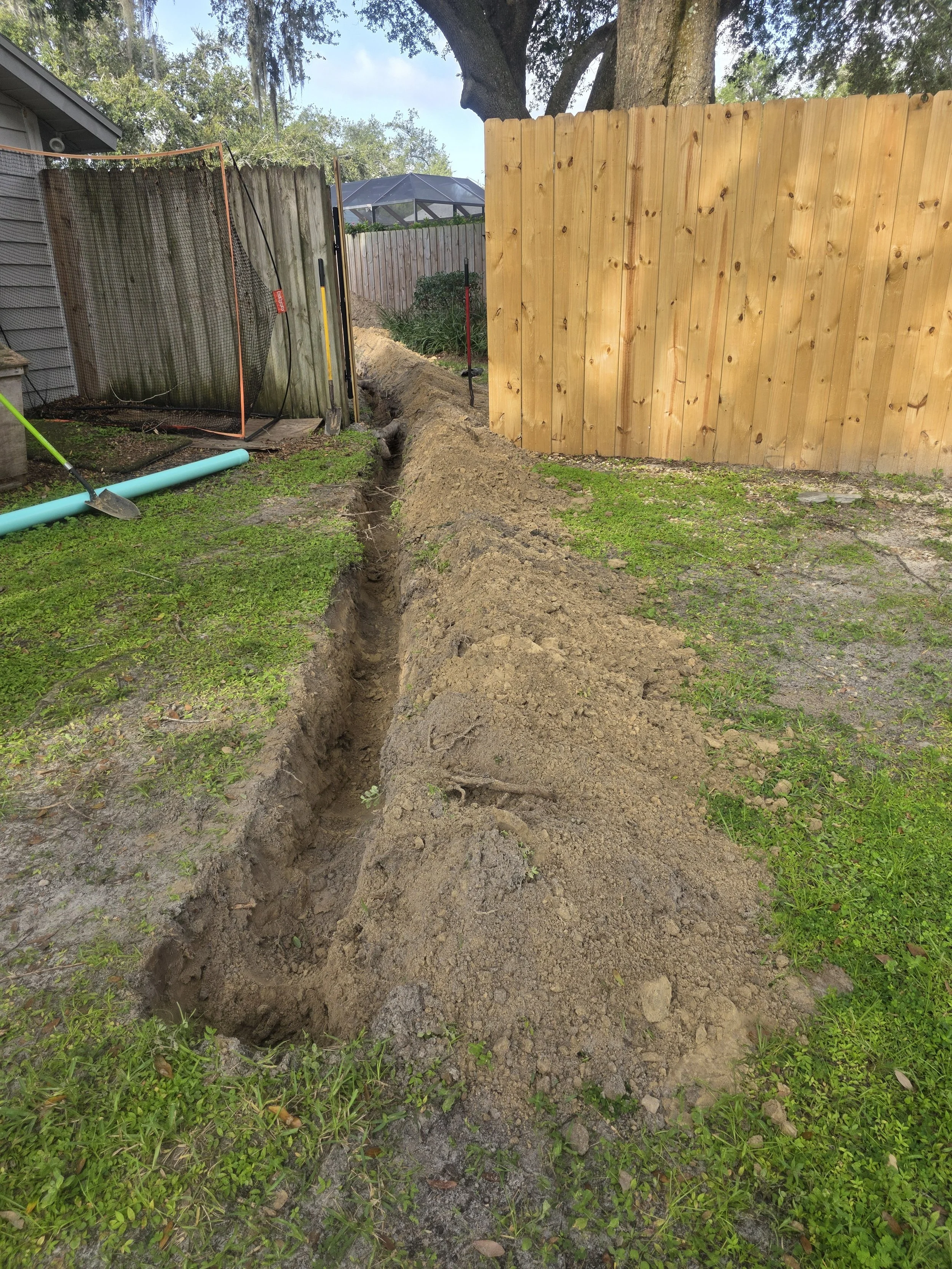 A backyard with a long trench dug in the ground, surrounded by grass and dirt. There is a wooden fence on the right and a chain-link gate on the left. Construction tools and materials are present, including a shovel and a large pipe, indicating ongoi