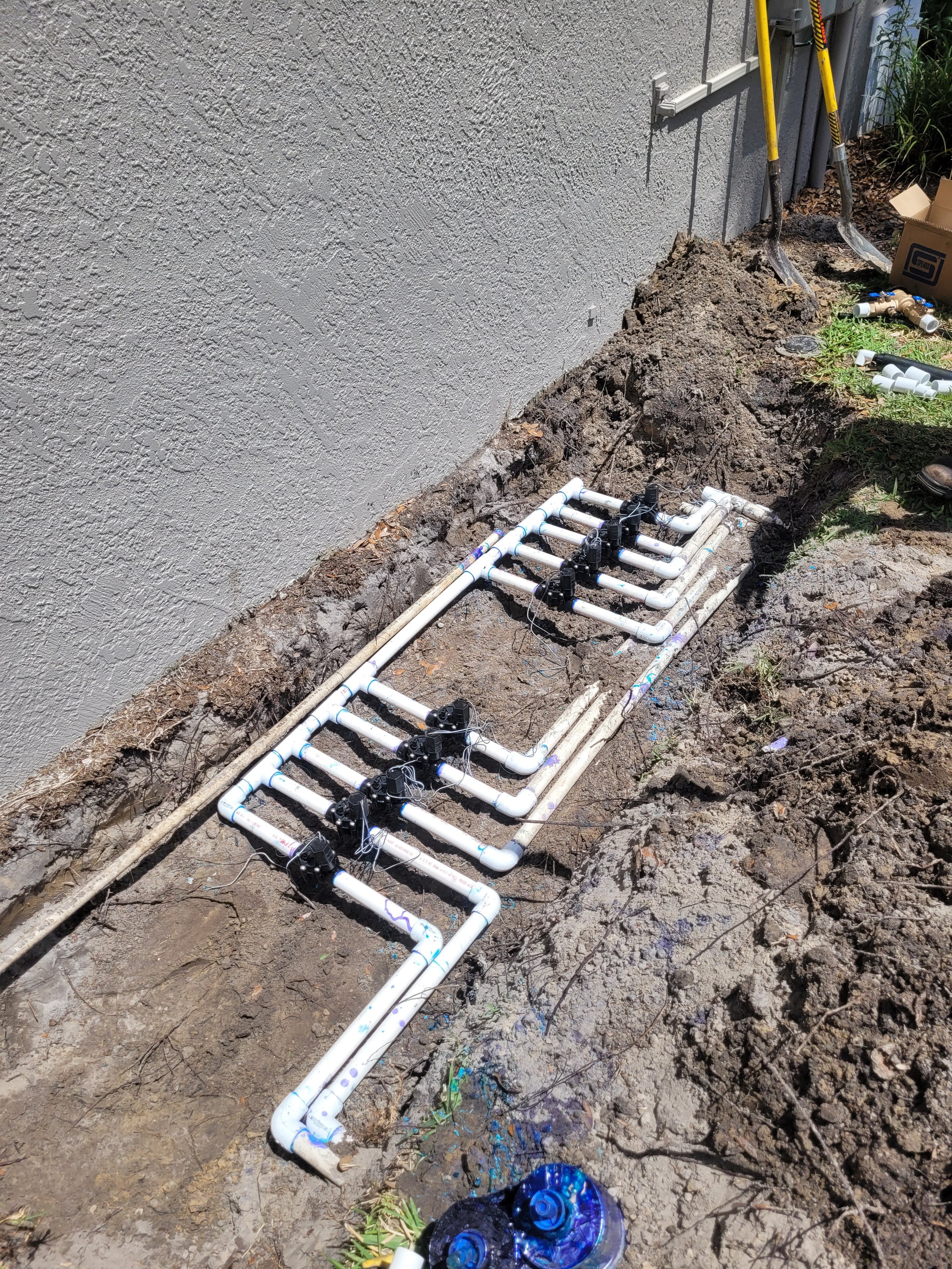 PVC pipe irrigation system installation in a dug trench beside a house wall, with tools and plumbing fittings nearby.