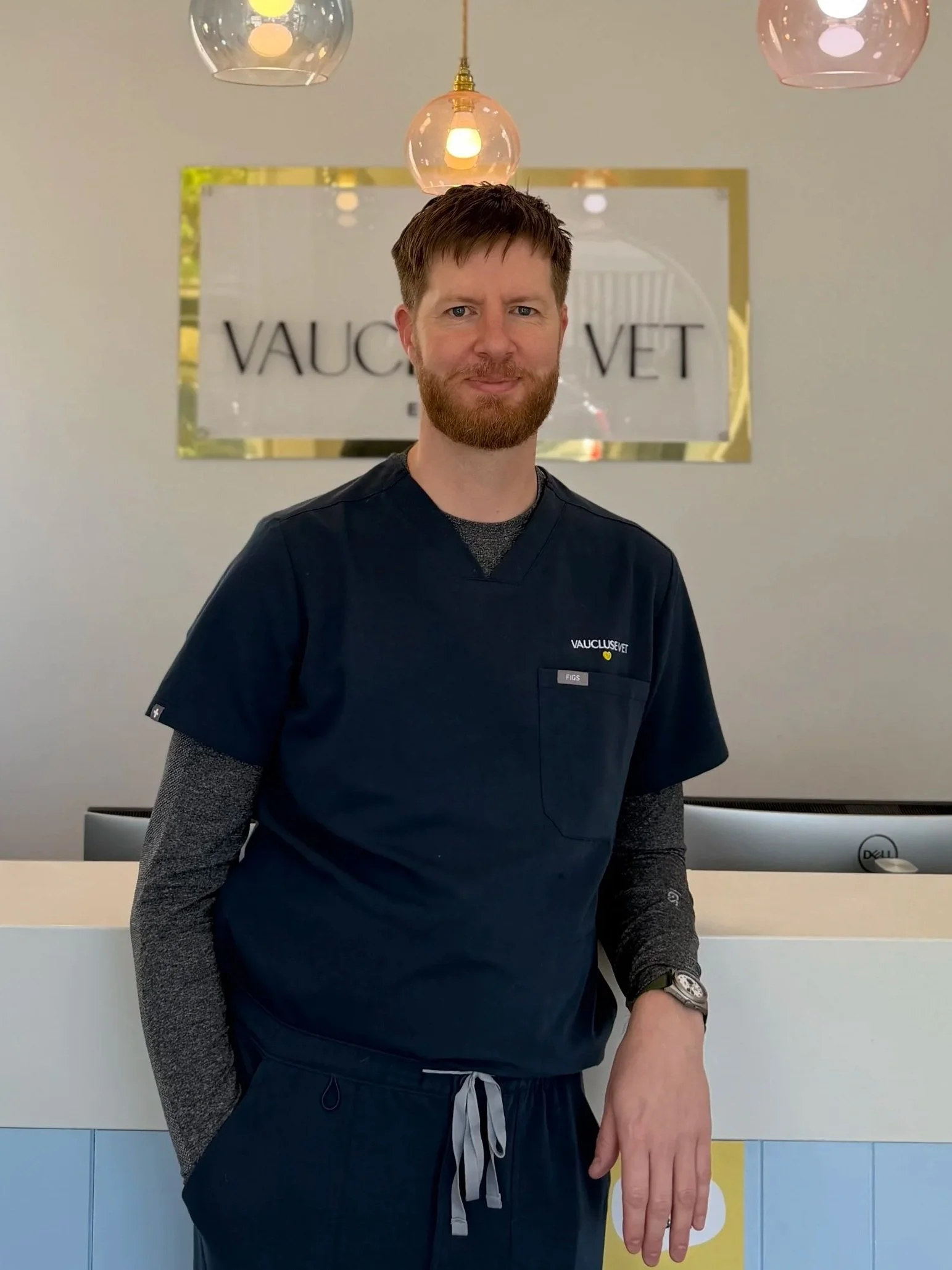 A man with a beard in medical scrubs standing at a reception desk in a veterinary clinic, with a sign that reads 'VAUCLUSE VET' in the background.