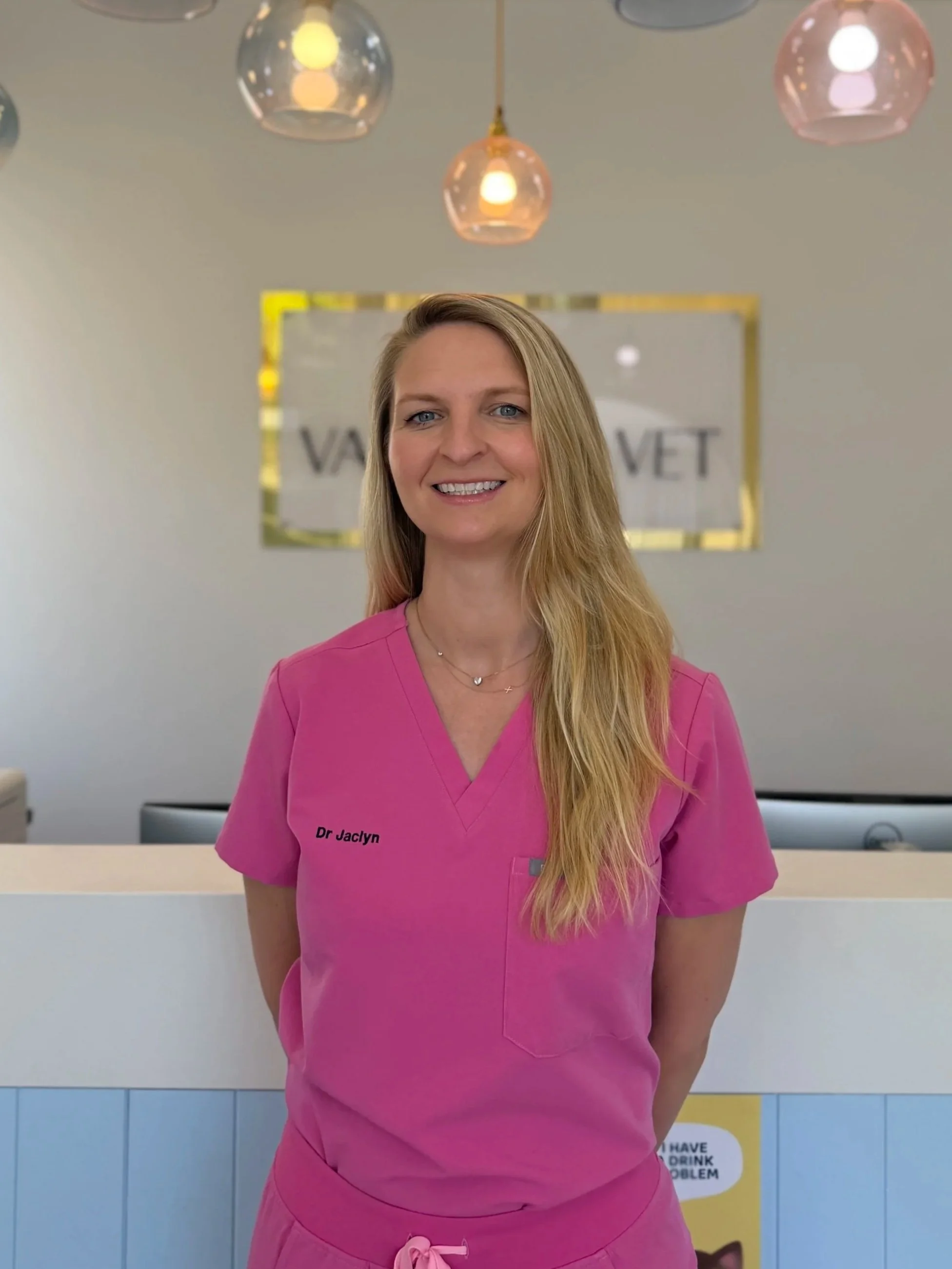 A smiling woman with long blonde hair in pink medical scrubs standing in a veterinary clinic.