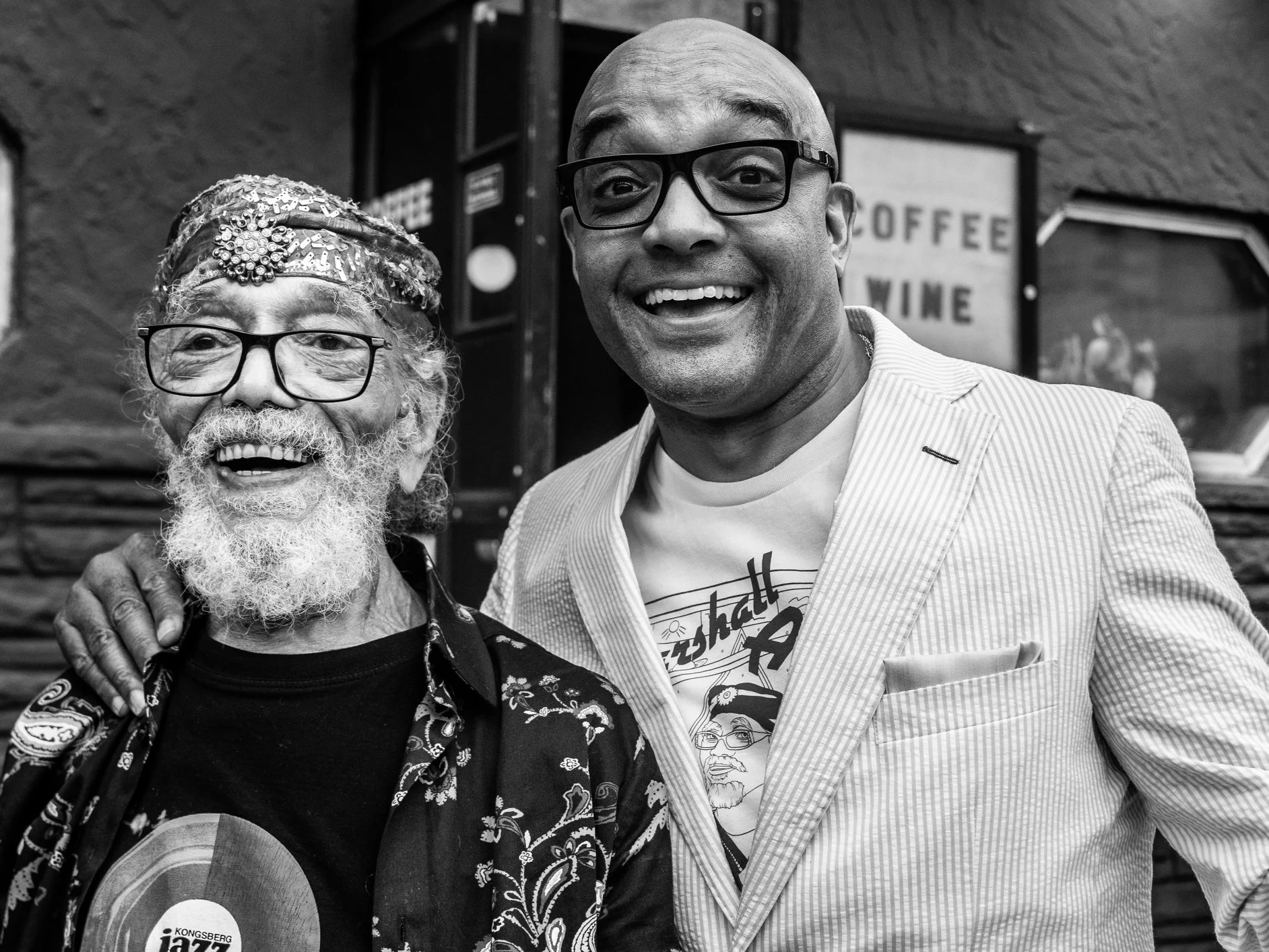 Two smiling men standing together, one elderly with a white beard, glasses, and wearing a hat and patterned shirt, the other middle-aged with glasses and a light striped blazer, in front of a sign that says 'Coffee' and 'Wine' in the background.