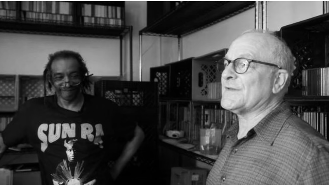 Two men standing in a room with shelves and equipment, one with dreadlocks and sunnies wearing a graphic t-shirt, the other with glasses wearing a checkered shirt.