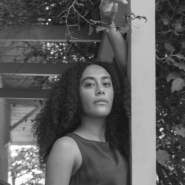 A woman with curly hair standing outdoors near a wooden structure and trees, with her arm raised and holding onto a branch.