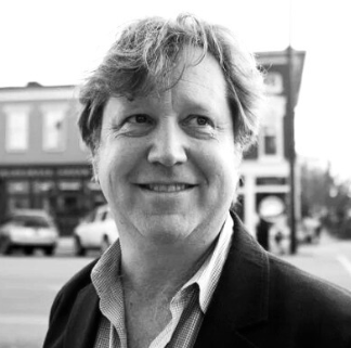 Black and white portrait of a smiling man with wavy hair, wearing a jacket and collared shirt, standing outdoors in an urban area.