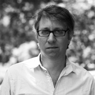 A man with glasses and short hair wearing a light-colored shirt, standing outdoors with blurred trees in the background.