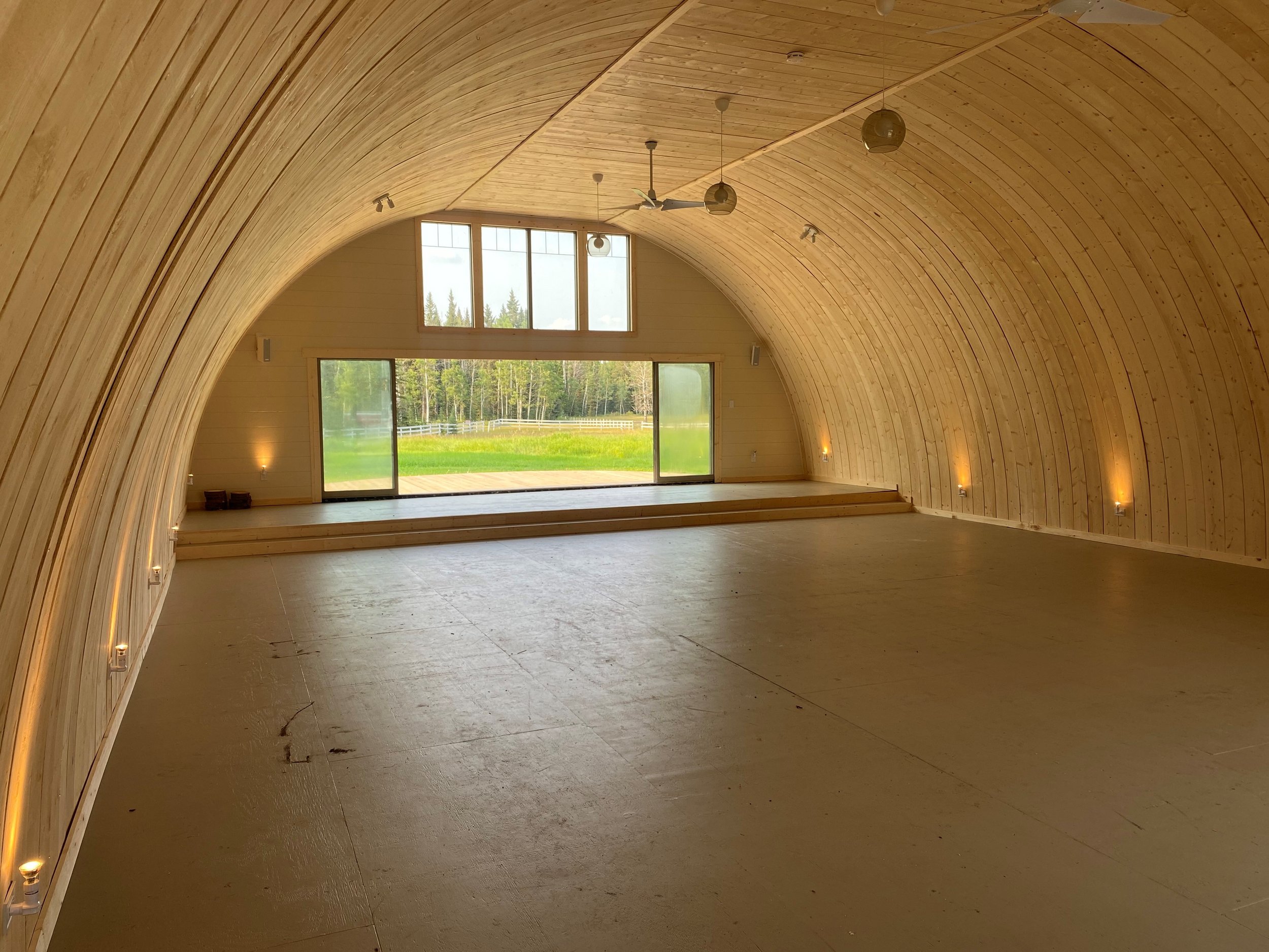 Empty indoor space with arched wooden ceiling, large windows and sliding glass doors revealing a green outdoor landscape, small steps leading to the outside, and minimal lighting fixtures along the walls.
