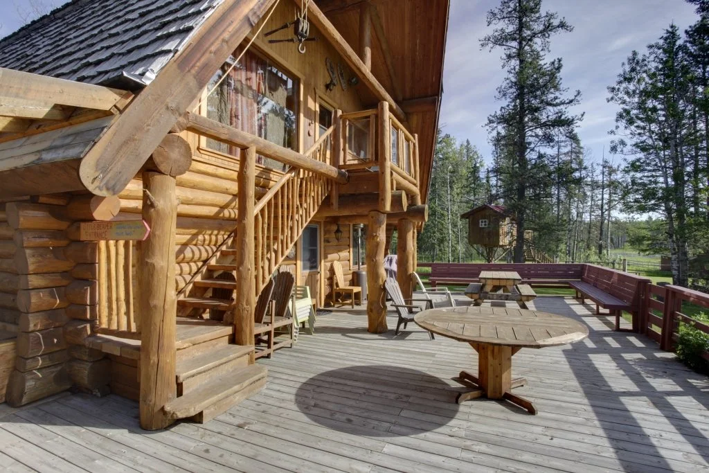 A wooden cabin with an outdoor deck featuring rustic furniture, surrounded by tall pine trees in a forested area.
