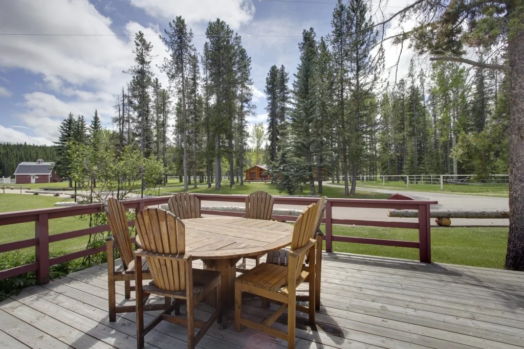 A wooden deck with a round table and six chairs, overlooking a grassy yard with trees in the background and a fence on the property.