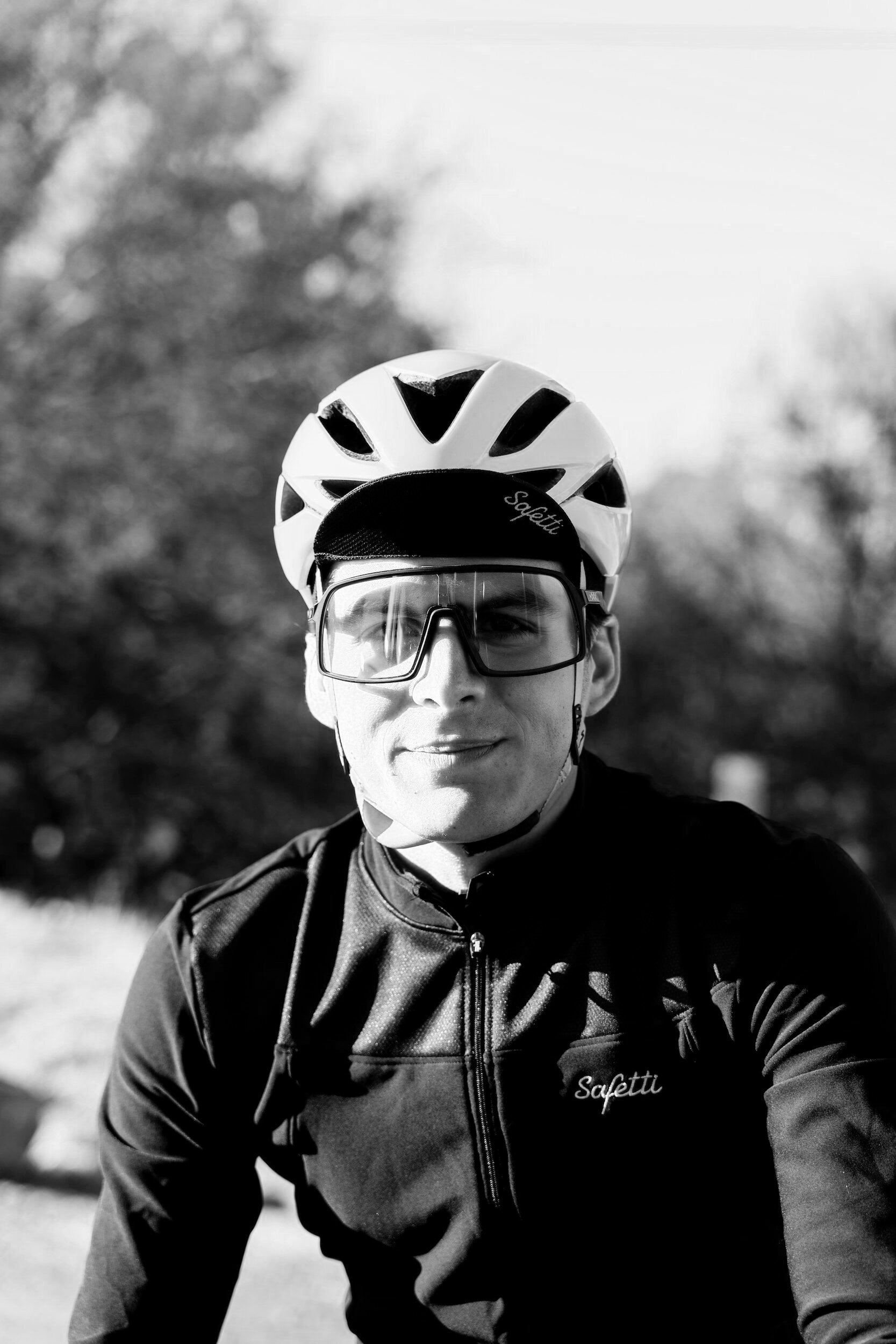 A cyclist wearing a helmet and sunglasses, smiling, outdoors with trees in the background.