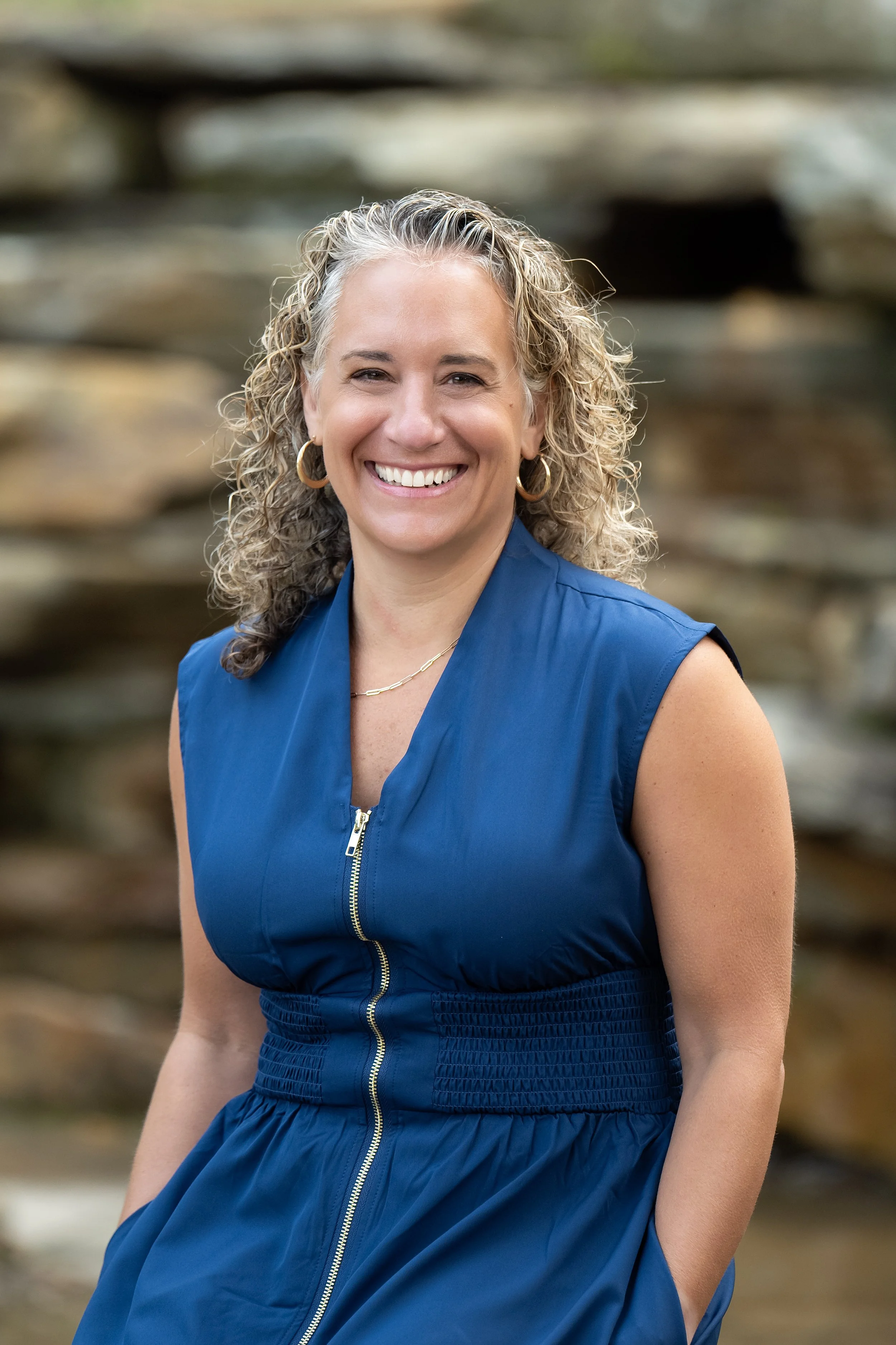 South Hills, Pittsburgh Psychologist. A woman with curly blonde hair smiling outdoors, wearing a blue sleeveless dress with a zipper.