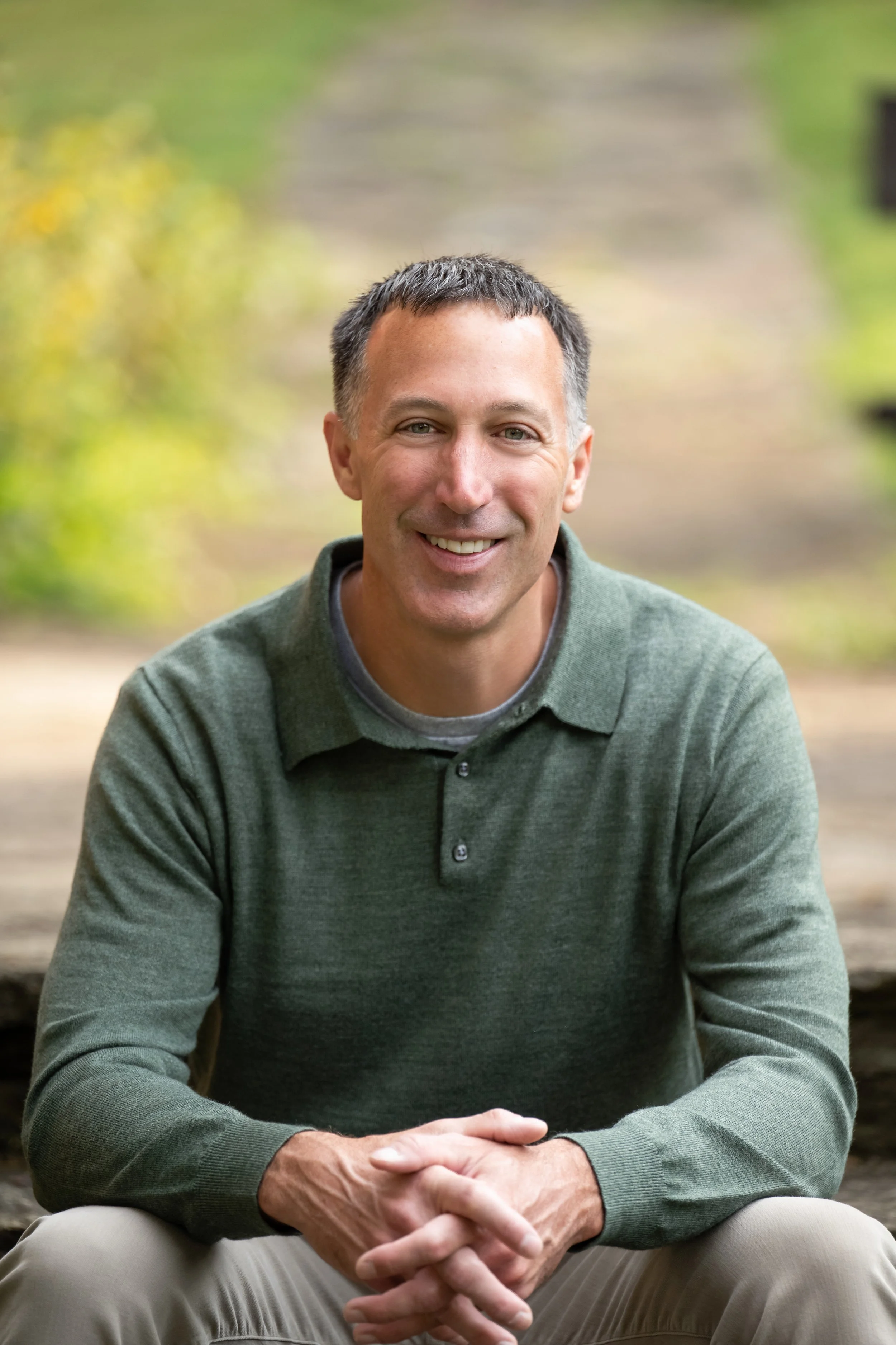 South Hills, Pittsburgh Psychologist.  A man smiling while sitting outdoors on a wooden surface, with a blurred background of greenery.