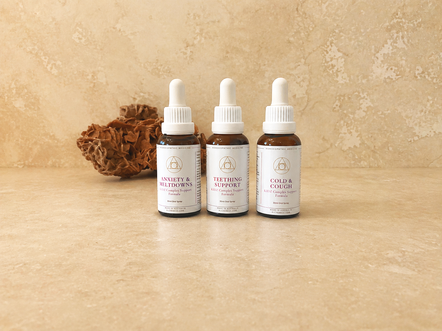 Four brown glass bottles of homeopathic liquid remedies with black droppers, labeled with names like Teething Support, Courage & Comfort, Anxiety + Meltdowns, and Sore Tummy, arranged on a white surface, with a small round stone beneath the first bottle.