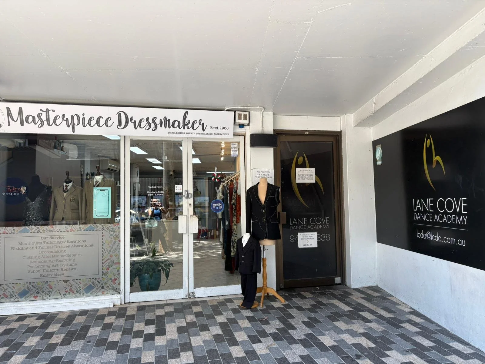 Storefront with a sign reading 'Masterpiece Dressmaker' and an entrance to Lane Cove Dance Academy. Mannequins display suits, and there are clothing items inside the store.