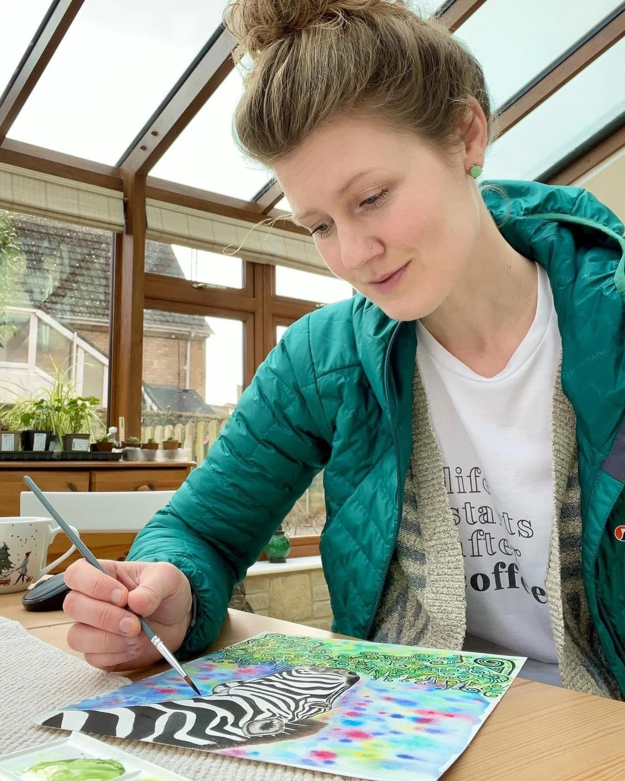 Woman painting a colorful, psychedelic zebra artwork in a sunlit greenhouse.