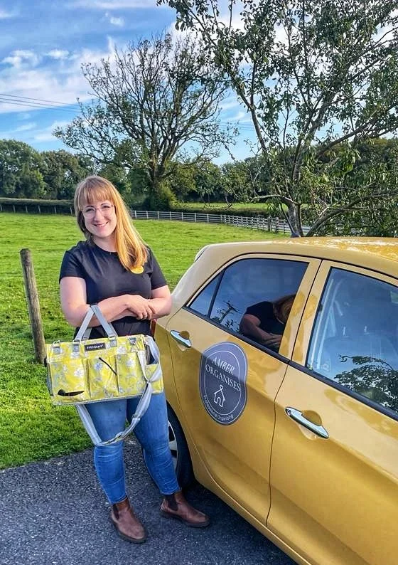 Amber wears a black shirt and jeans, standing outdoors next to a yellow car with a 'Amber Organises' sticker on the door, holding a yellow patterned bag, in a grassy area with trees and a wooden fence in the background.