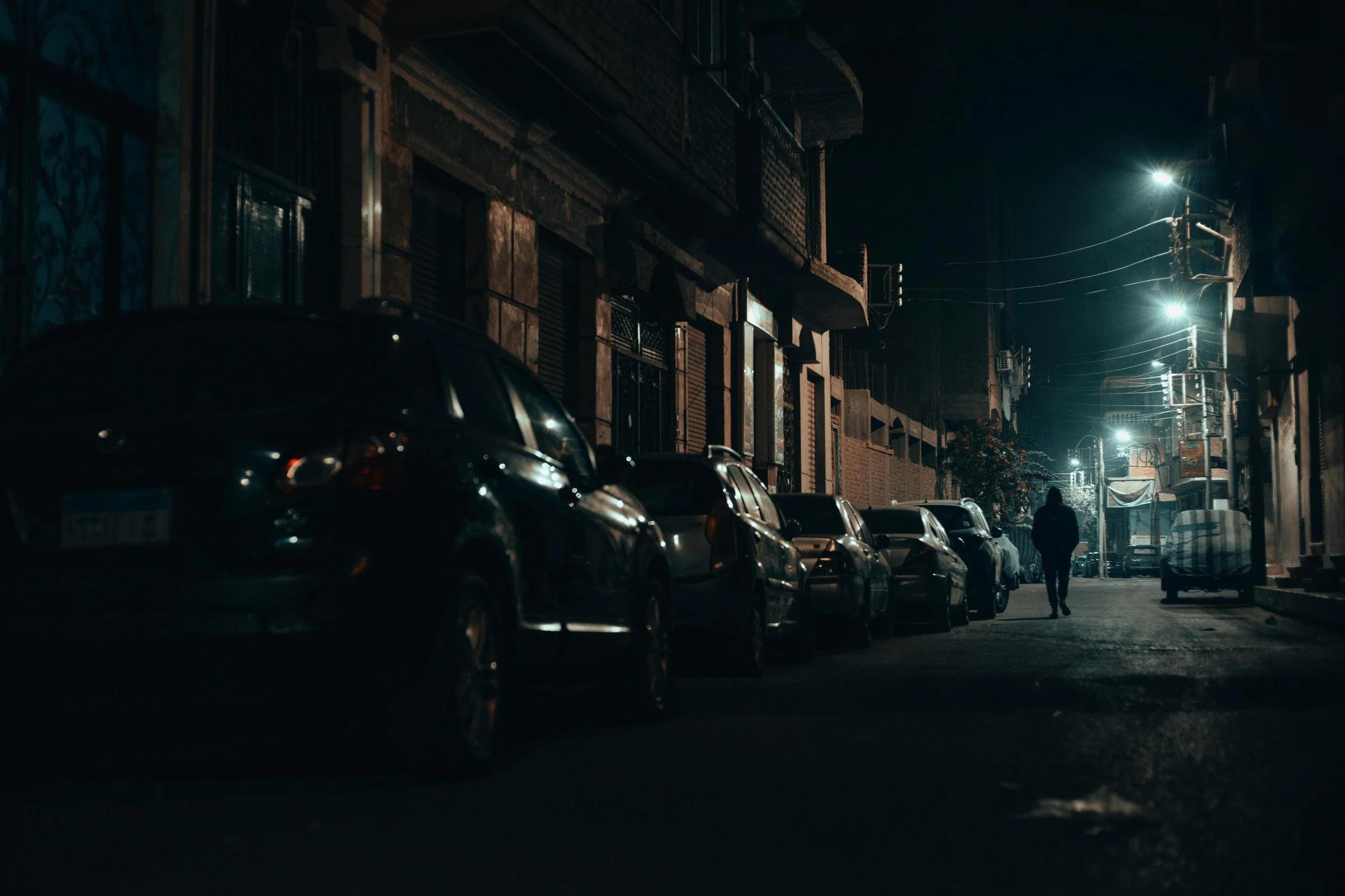 Nighttime street scene with parked cars along a dimly lit urban street. A person walks down the sidewalk. Buildings with closed shutters line the street, and streetlights illuminate the scene.