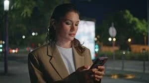 Woman using her phone at night in a city setting, representing personal safety awareness, staying connected and using mobile tools for safe travel and event journeys.
