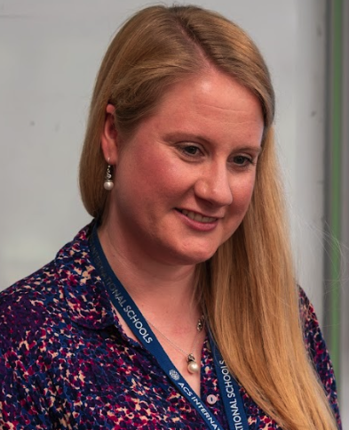 A woman with long blonde hair wearing a patterned blouse and a lanyard, smiling.
