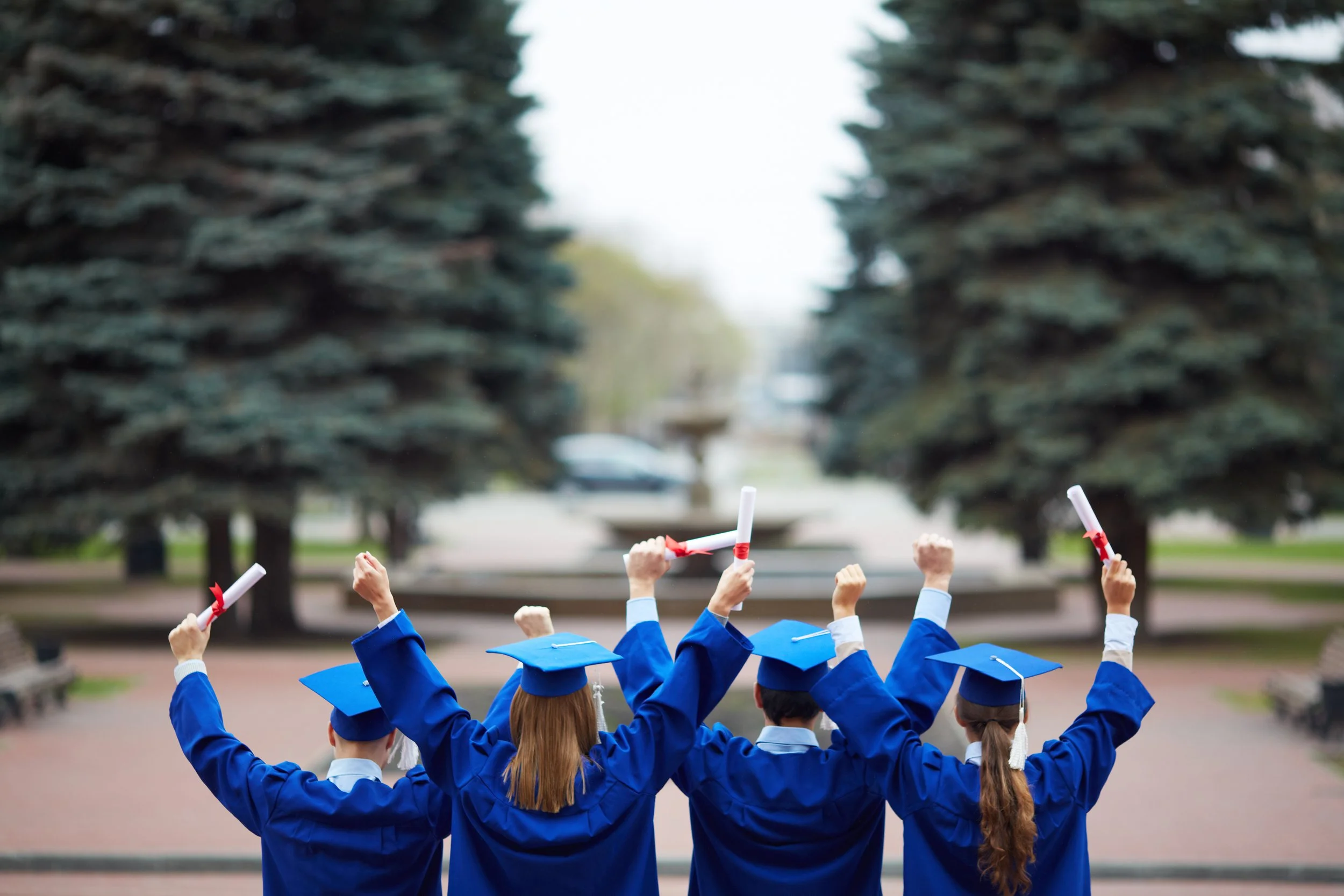 Group of graduates in blue gowns and caps holding diplomas, celebrating outdoors.