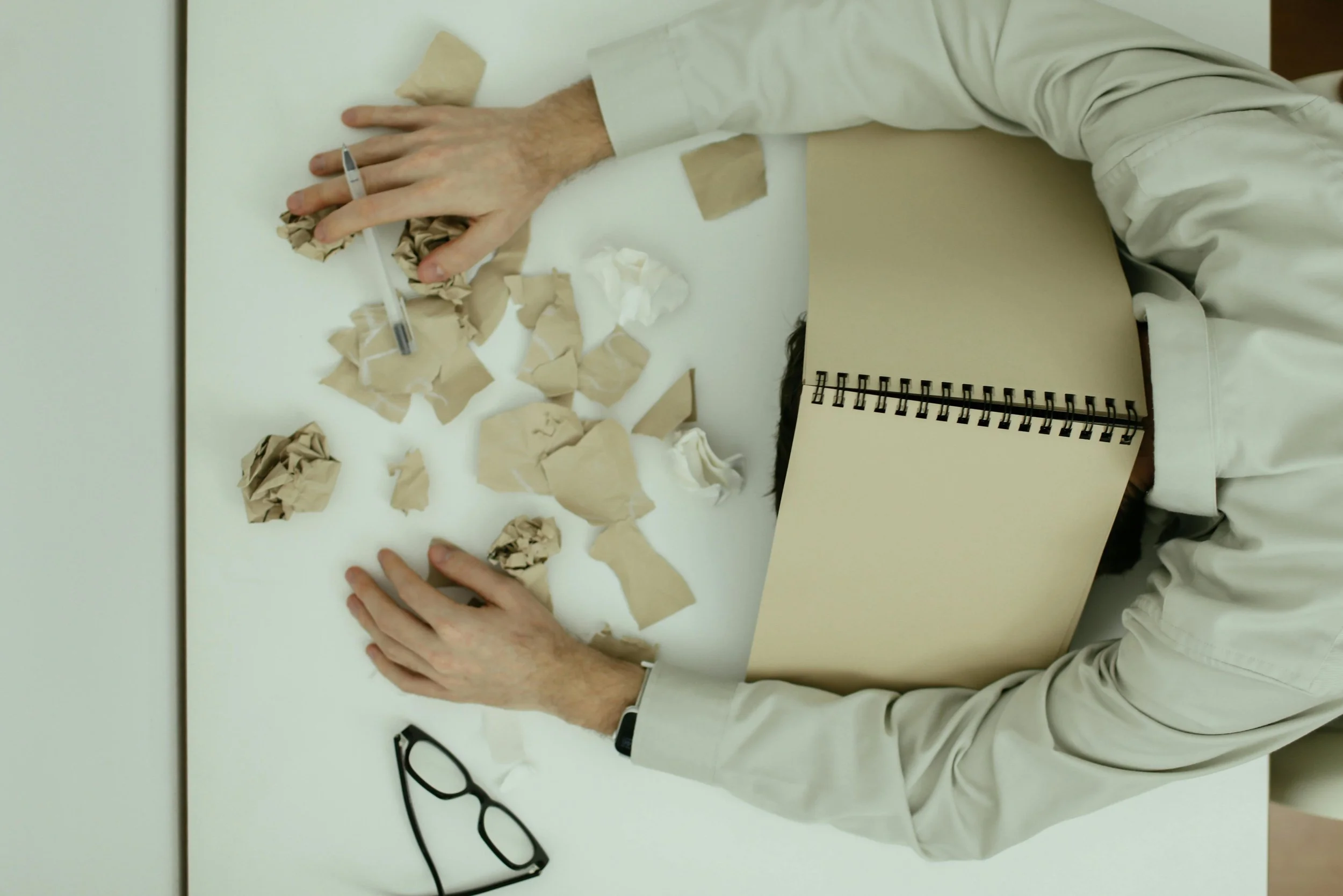 Person overwhelmed at a desk with head down on a notebook, surrounded by crumpled paper, representing stress, mental exhaustion and feeling unable to cope.