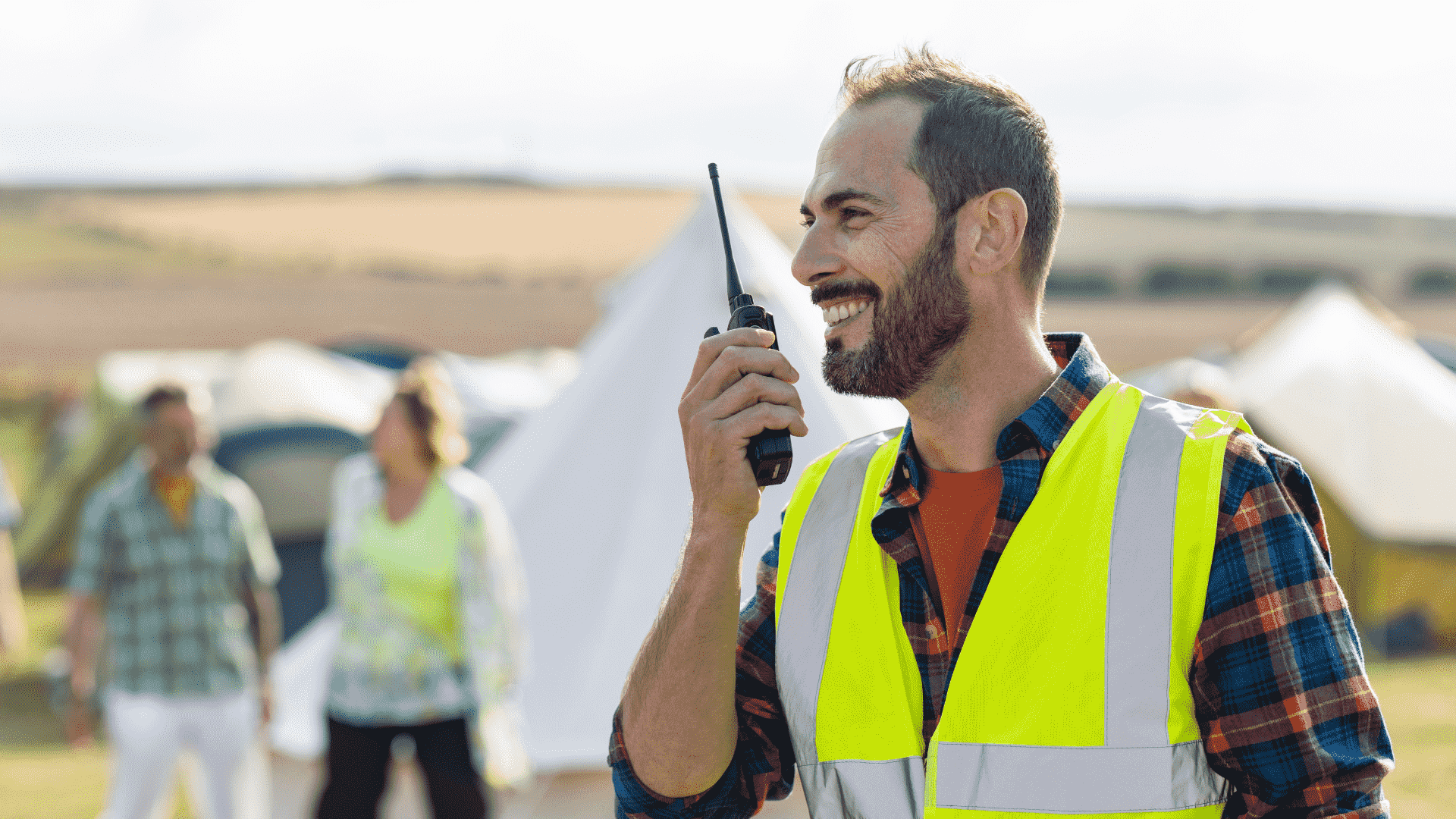 A smiling festival steward wearing a high-visibility vest and speaking into a radio, representing the on-site security, welfare and support services festivalgoers can turn to for help, alongside trusted safety information available through imabi.