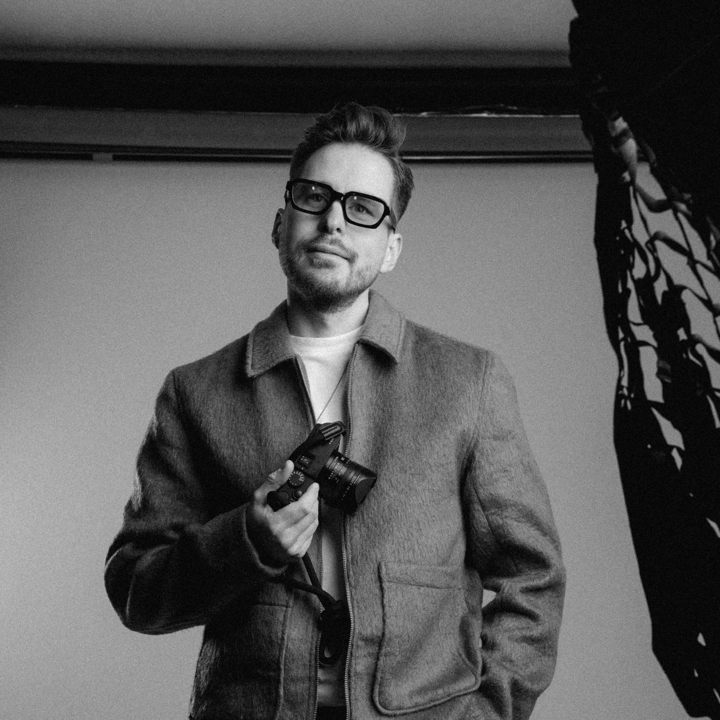 Schwarz-weiß-Foto eines Mannes mit Brille, der eine Kamera in der Hand hält und in die Kamera schaut.