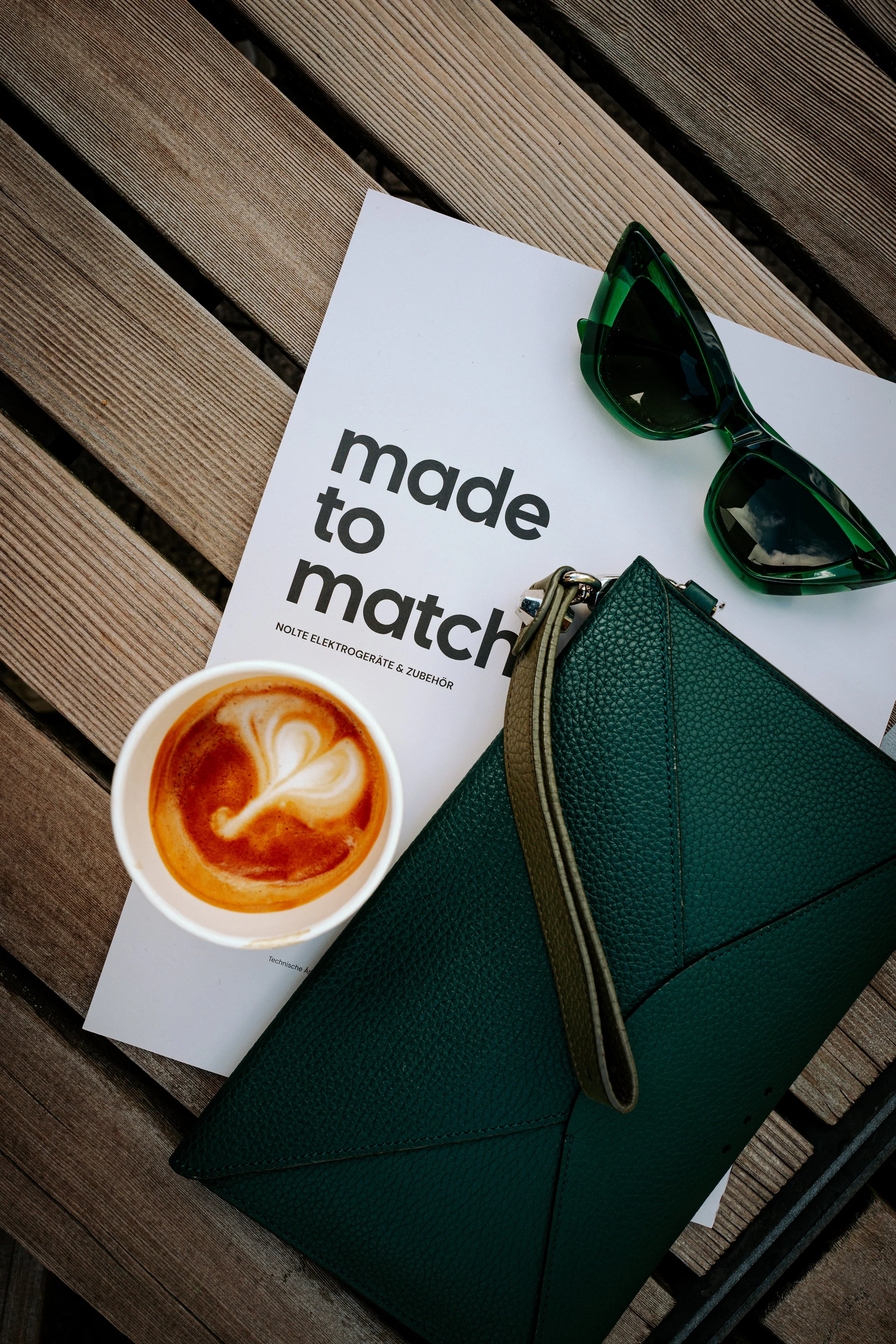 Lifestyle-Produktfoto: Flat White, Sonnenbrille und grüne Ledertasche auf Holzdeck