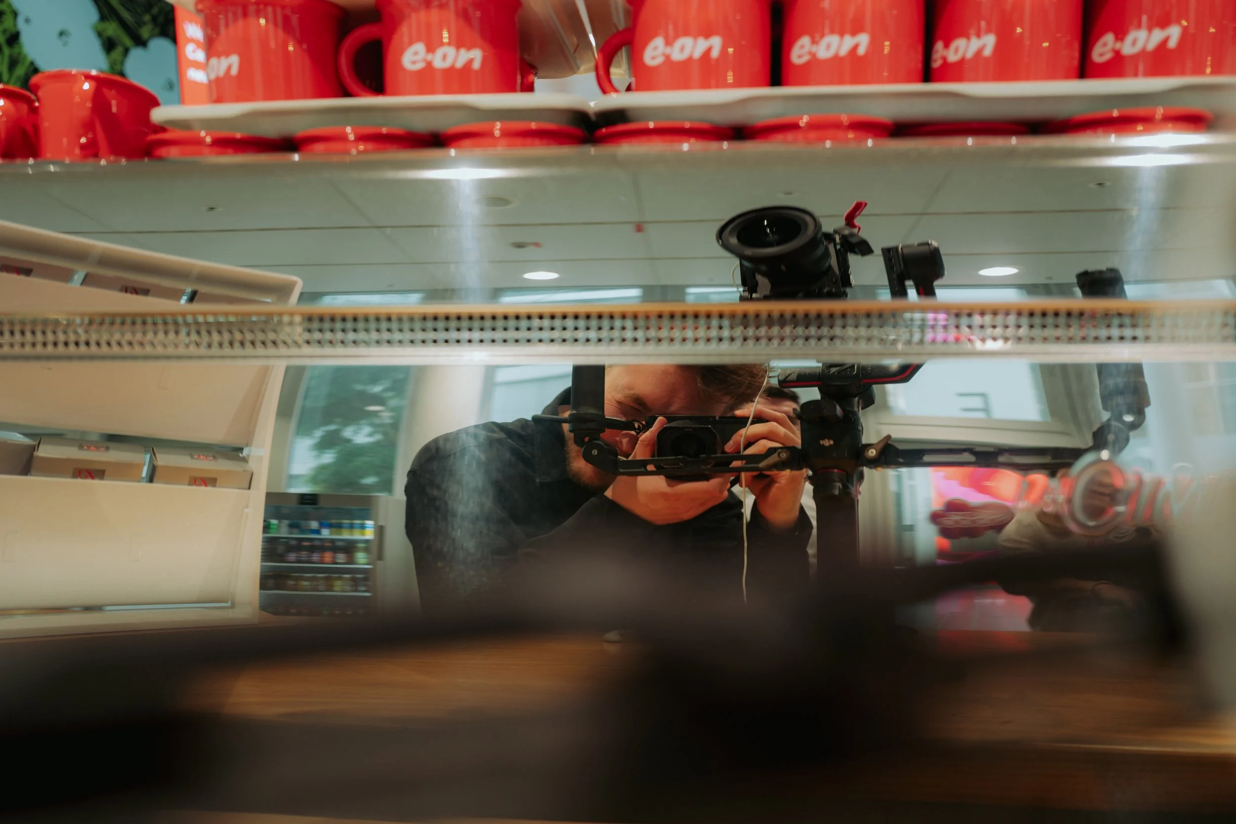Spiegelbild eines Fotografen, der in einem Supermarkt mit roten Eon-Kaffeetassen auf einem Regal fotografiert.