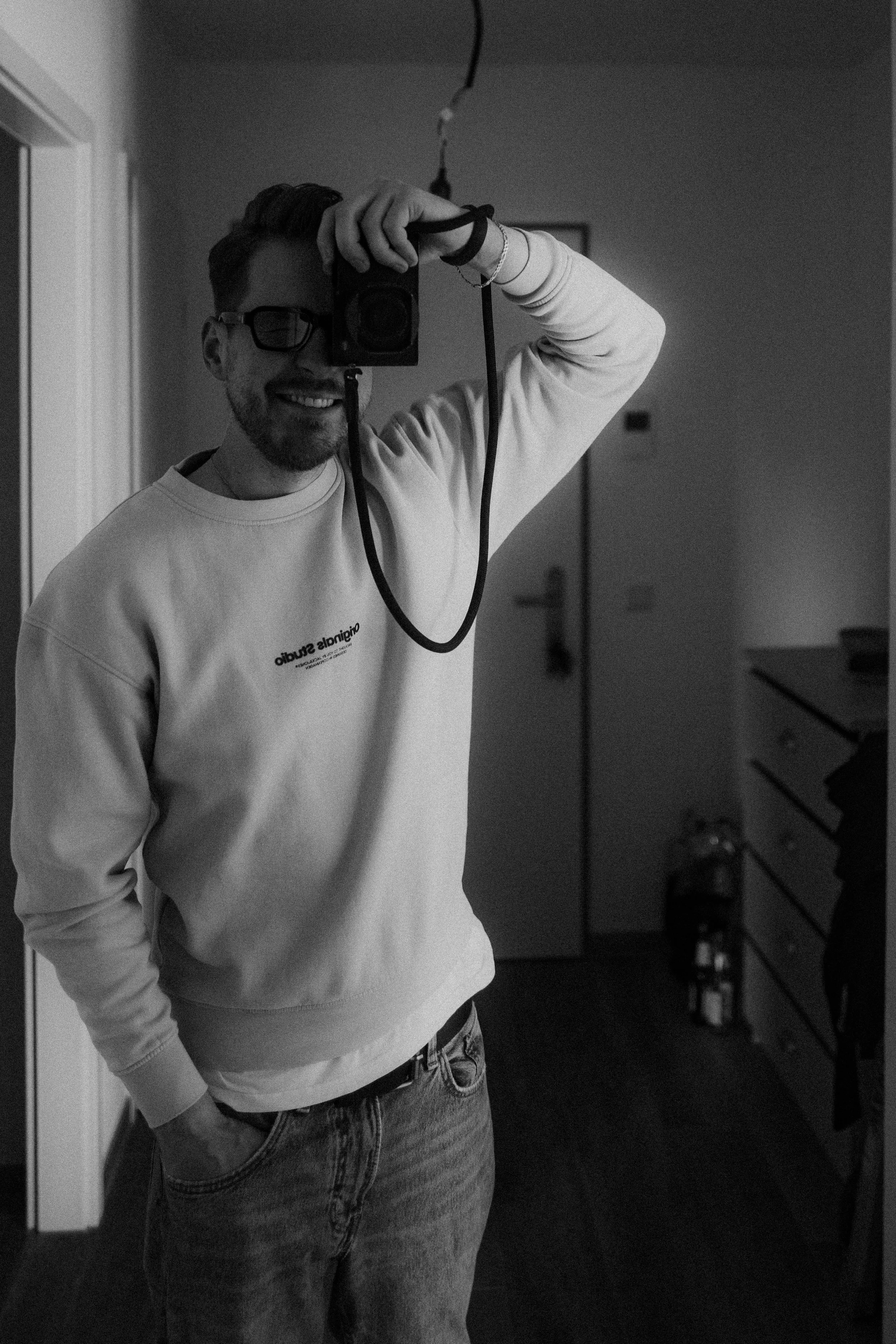 Junger Mann macht ein Selfie im Spiegel, trägt Brille und eine Sweatshirt, lächelt und posiert in einem Raum, schwarz-weiß Aufnahme.