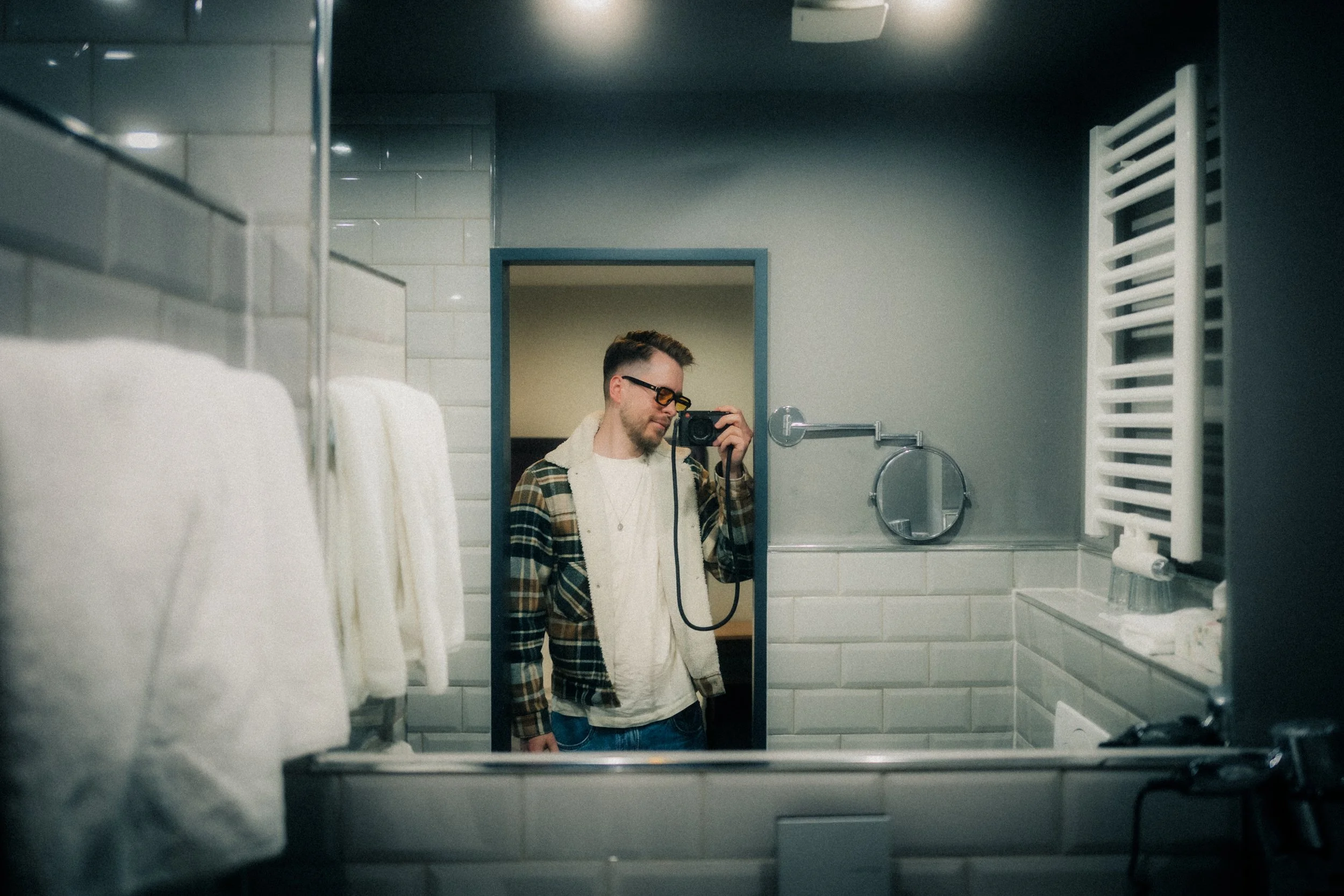 Junger Mann fotografiert sich selbst im Spiegel eines Badezimmers, trägt eine kariert Jacke und Brille.