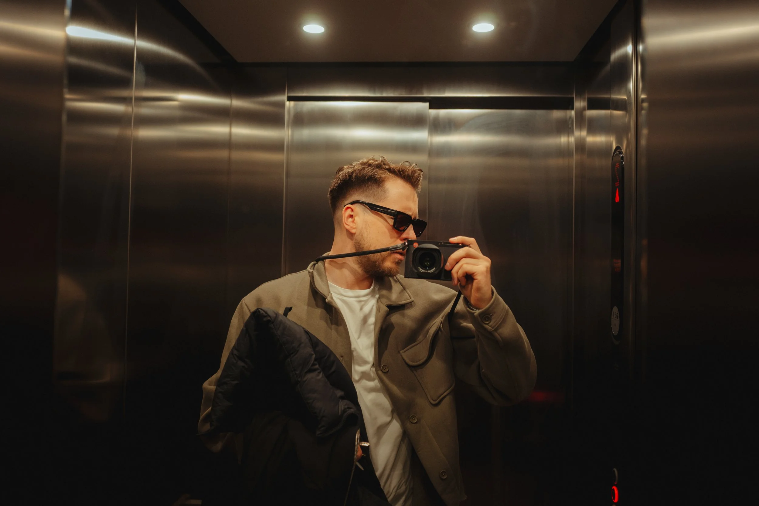 Ein junger Mann in einem Aufzug, trägt Sonnenbrille und hält eine Kamera, trägt eine Jacke über der Schulter.