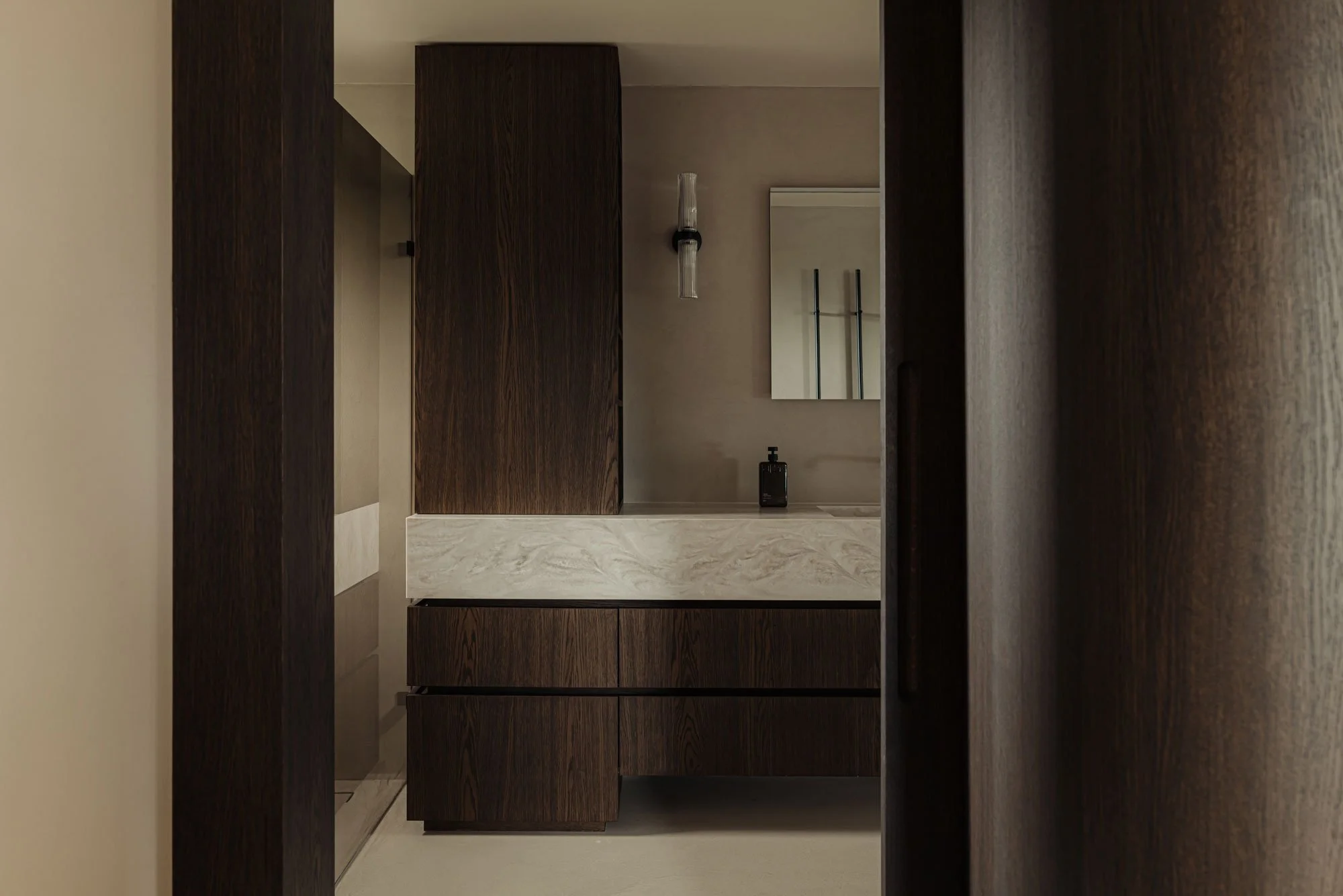 Doorkijk vanuit de master bedroom naar een high-end badkamer en suite met een luxe maatwerk wastafelmeubel van natuursteen en donker hout, ontworpen door Studio Engelhard uit Amsterdam.