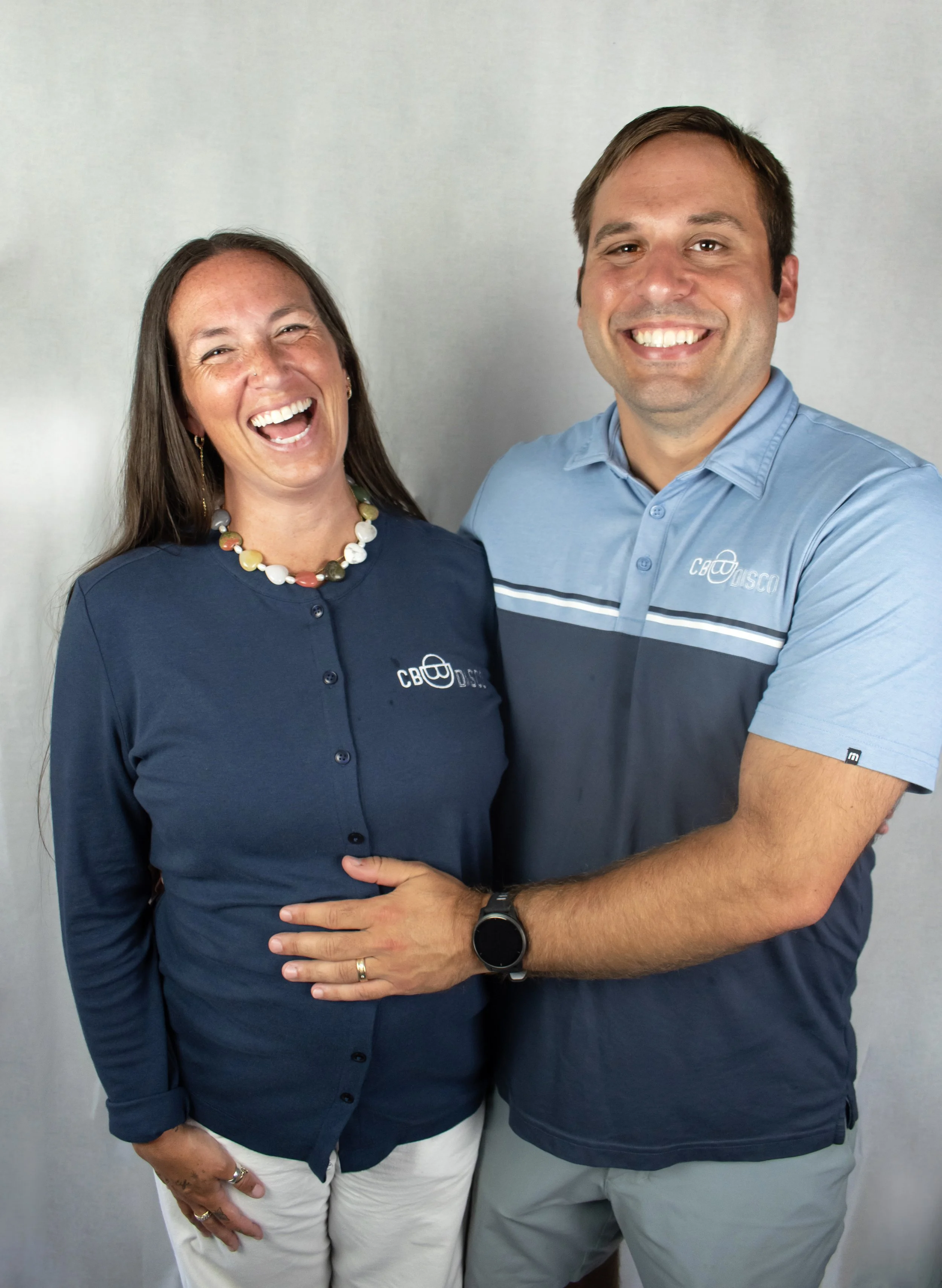 A man and woman smiling and posing together, the woman is wearing a navy blue shirt with a company logo, and the man is wearing a blue polo shirt with the same logo, in front of a plain white wall.