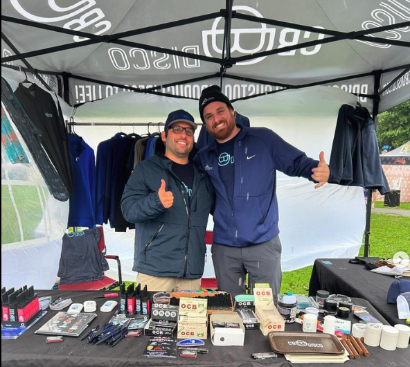 Two men smiling and giving thumbs up at an outdoor booth with various products displayed on the table, under a black canopy tent.