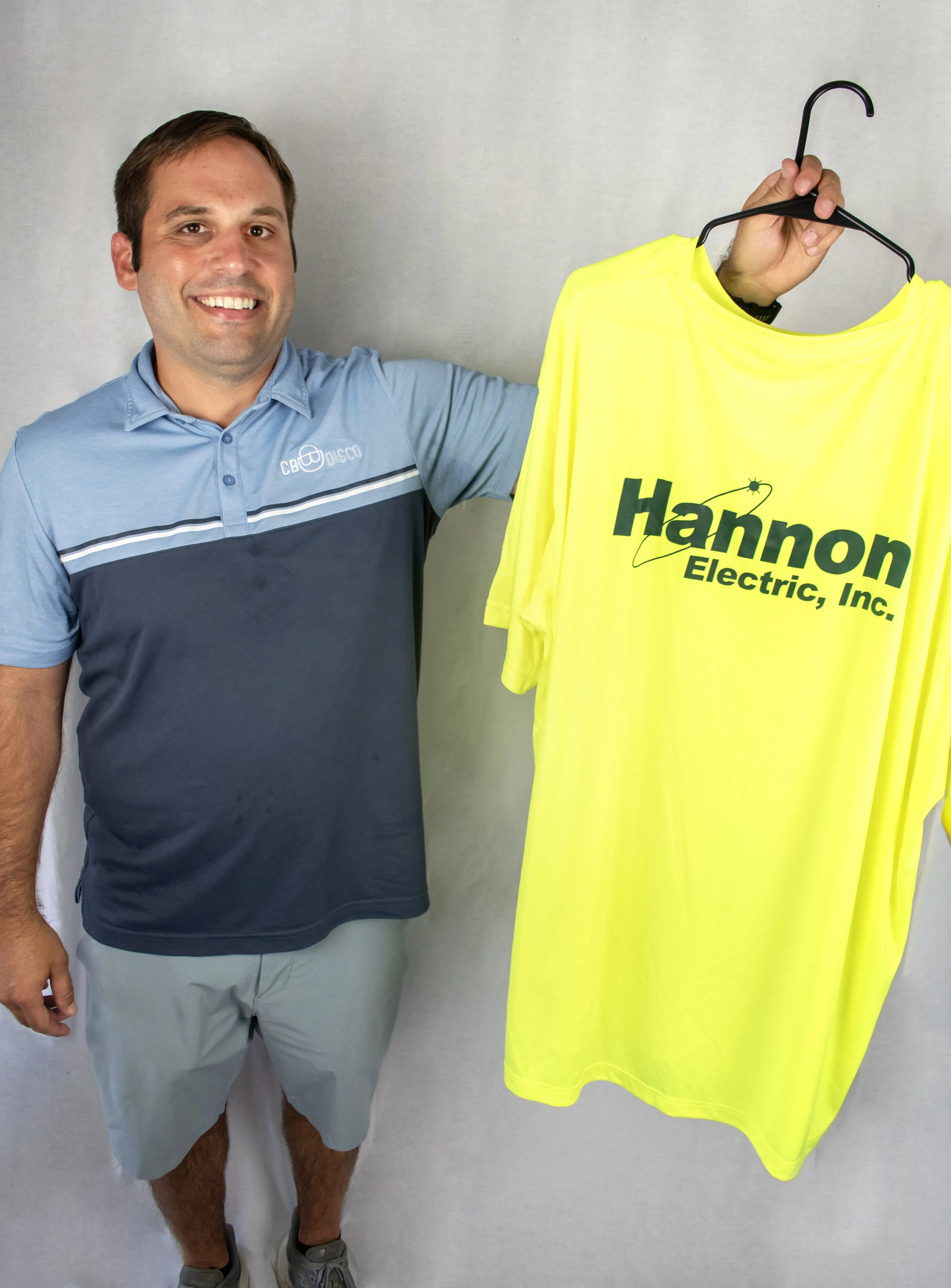 A man smiling and holding a yellow T-shirt with the logo 'Hannon Electric, Inc.' printed on it.